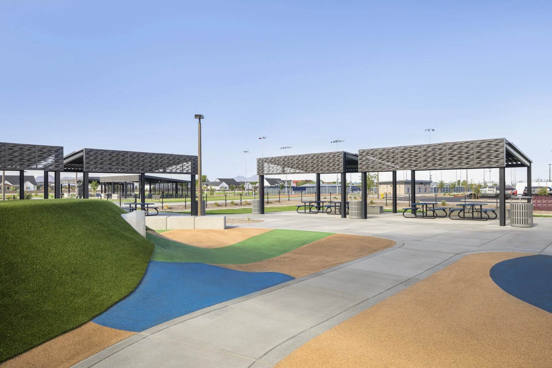 Empty outdoor pavilion with picnic tables at a park, colorful rubber playground surfacing, grassy hill, and a sports field in the background.