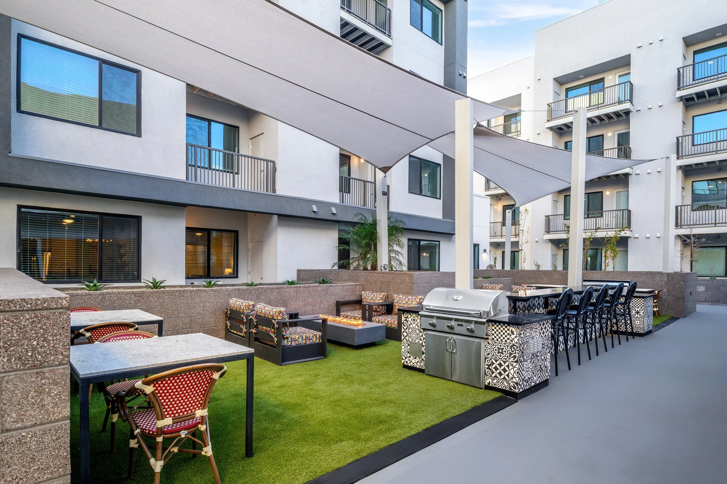 Outdoor courtyard with patio furniture, including tables, chairs, a fireplace, a grill, and shaded canopy, surrounded by modern apartment buildings.