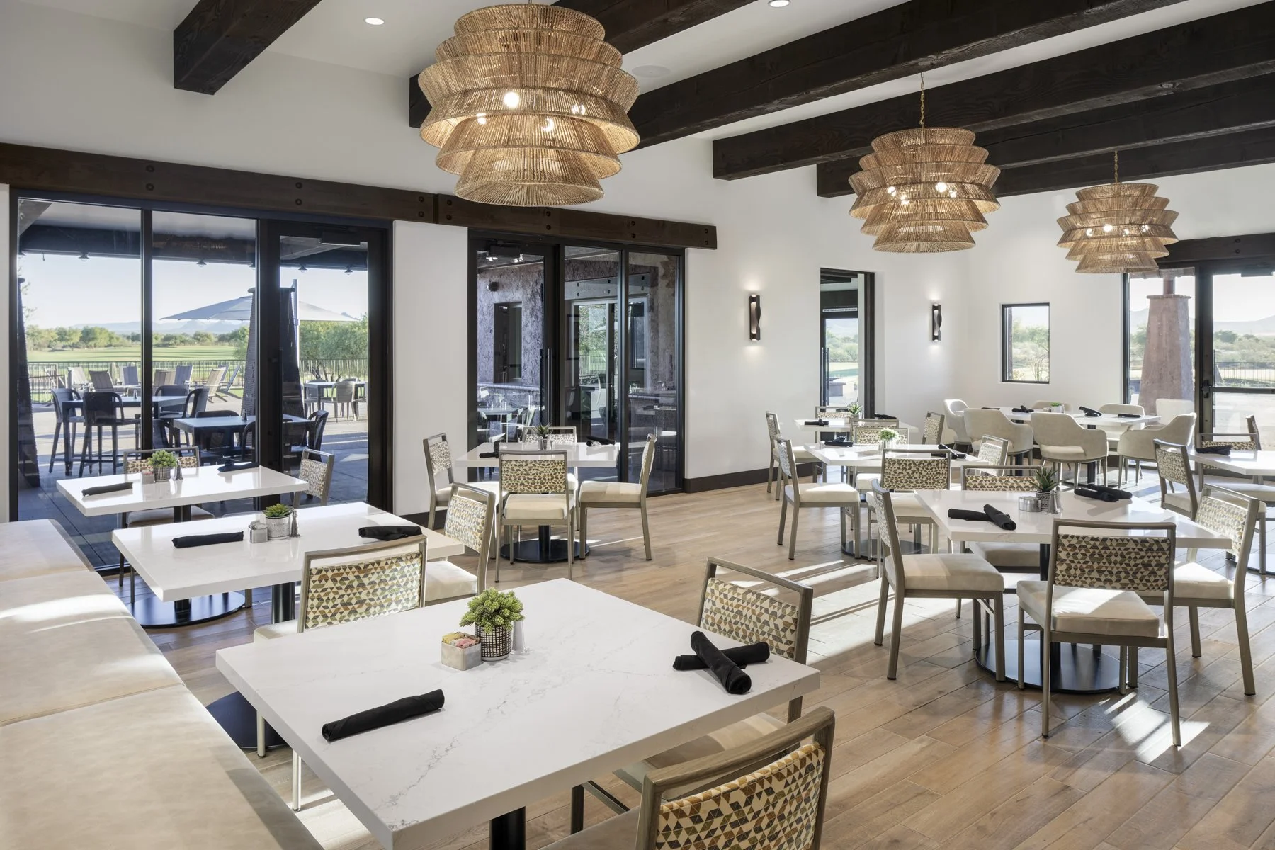 A bright dining area with white marble tables, beige cushioned chairs, and large woven pendant lights hanging from a ceiling with dark wooden beams. Large sliding glass doors and windows let in natural light and offer views of an outdoor patio and gr