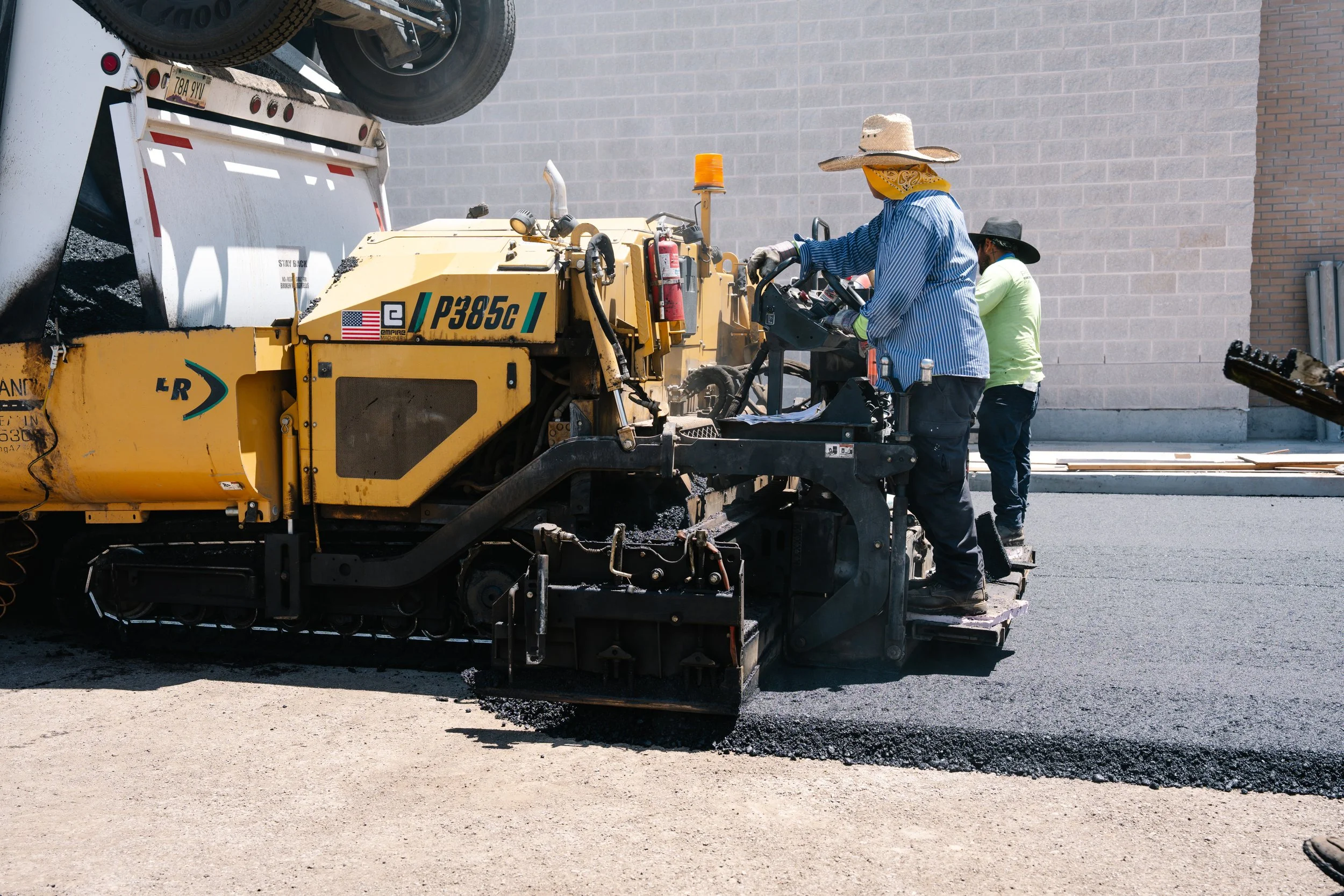 Two construction workers operating a yellow asphalt paving machine on a new road, with one worker in a blue striped shirt and straw hat, and another in a yellow shirt and black hat, against a building with a brick wall.