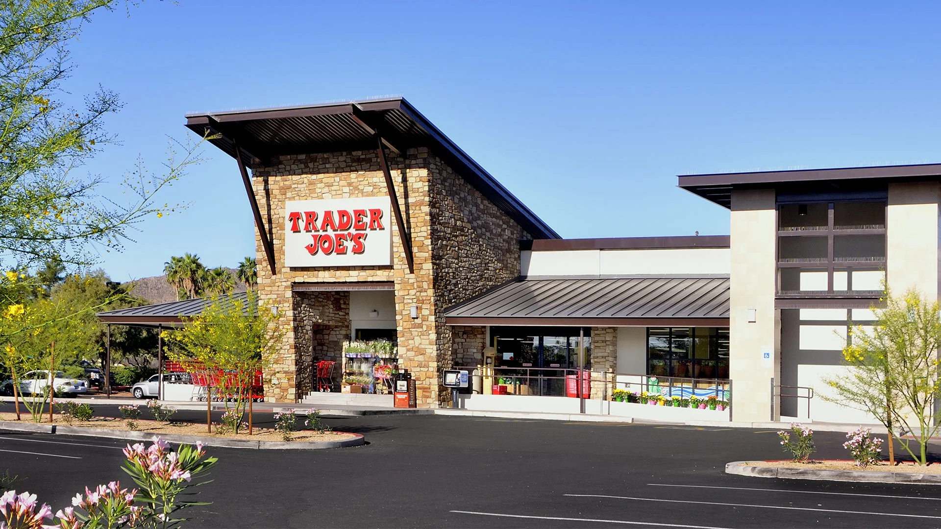 Exterior view of a Trader Joe's grocery store with a stone facade, signage, parking lot, and landscaping with flowering trees and plants.