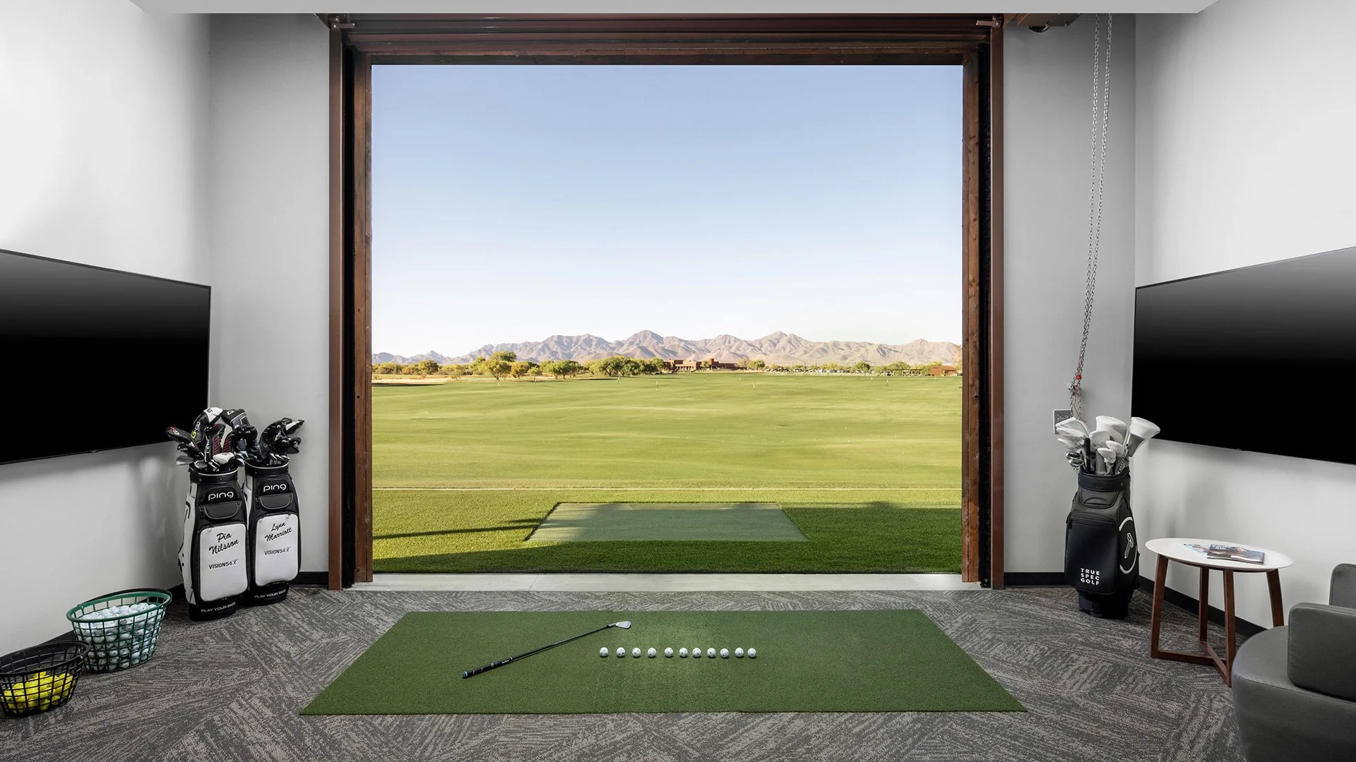 Indoor golf practice room with a view of a golf course and mountains. Contains golf clubs in bags, a small round table, and a hook with golf balls on the ceiling.