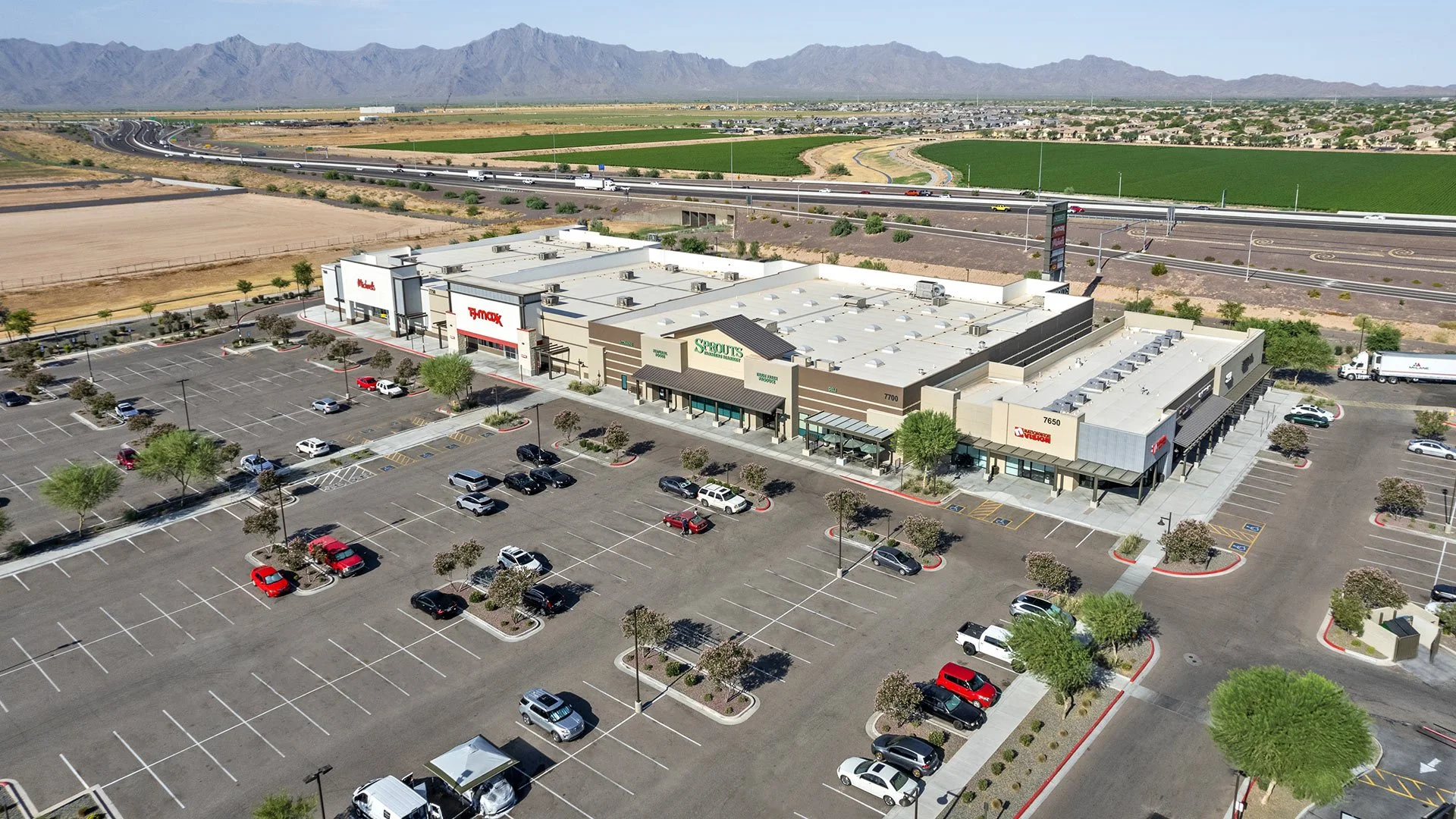 Large shopping center with parking lot and trees, featuring stores such as Sprouts, Ross, Bed Bath & Beyond, and Vision. Mountain range in the background with highways and fields.