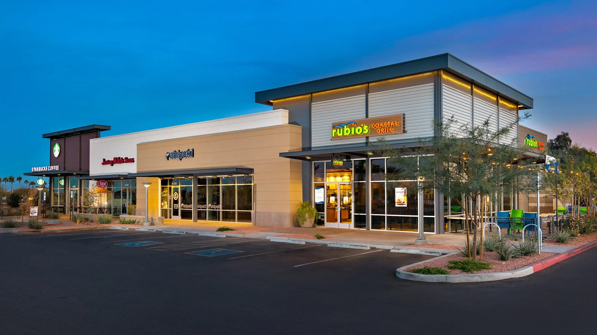 A modern shopping center with various storefronts including Starbucks Coffee, Jack in the Box, ahipoki, and Rubios Coastal Grill, outdoor seating, and a parking lot at dusk.