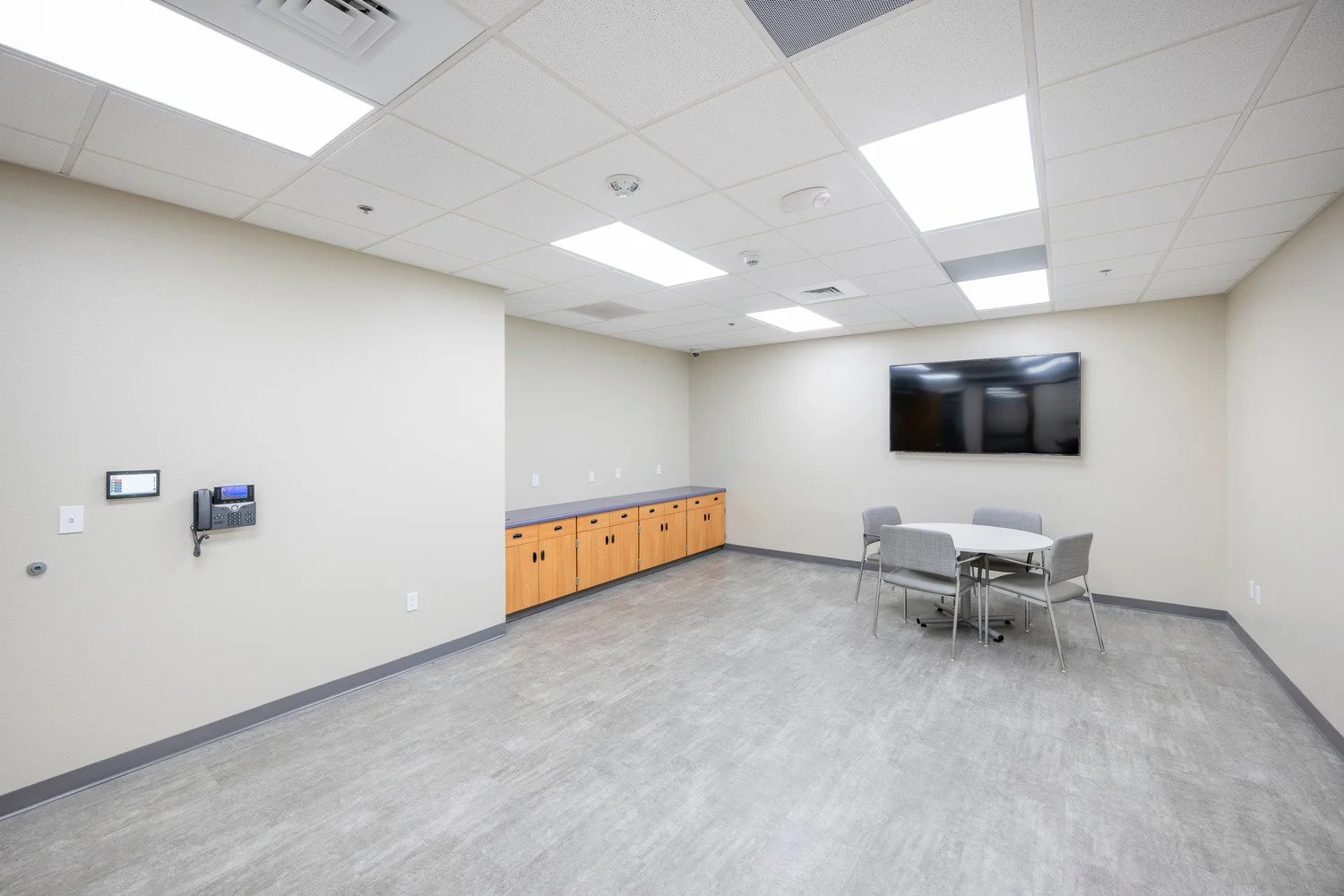 Empty conference room with a wall-mounted TV, a small round table with four chairs, built-in cabinets, and communication devices on the wall.
