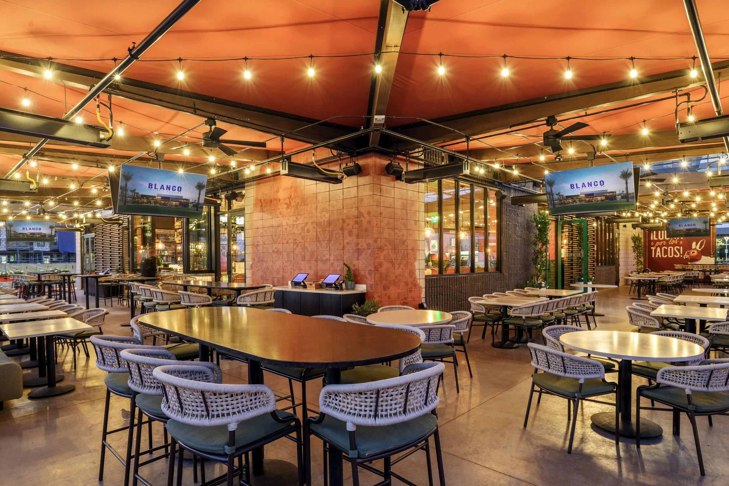 Empty restaurant or bar with round and rectangular tables, chairs, warm ambient lighting, TVs showing "BLANCO," and decorative plants.