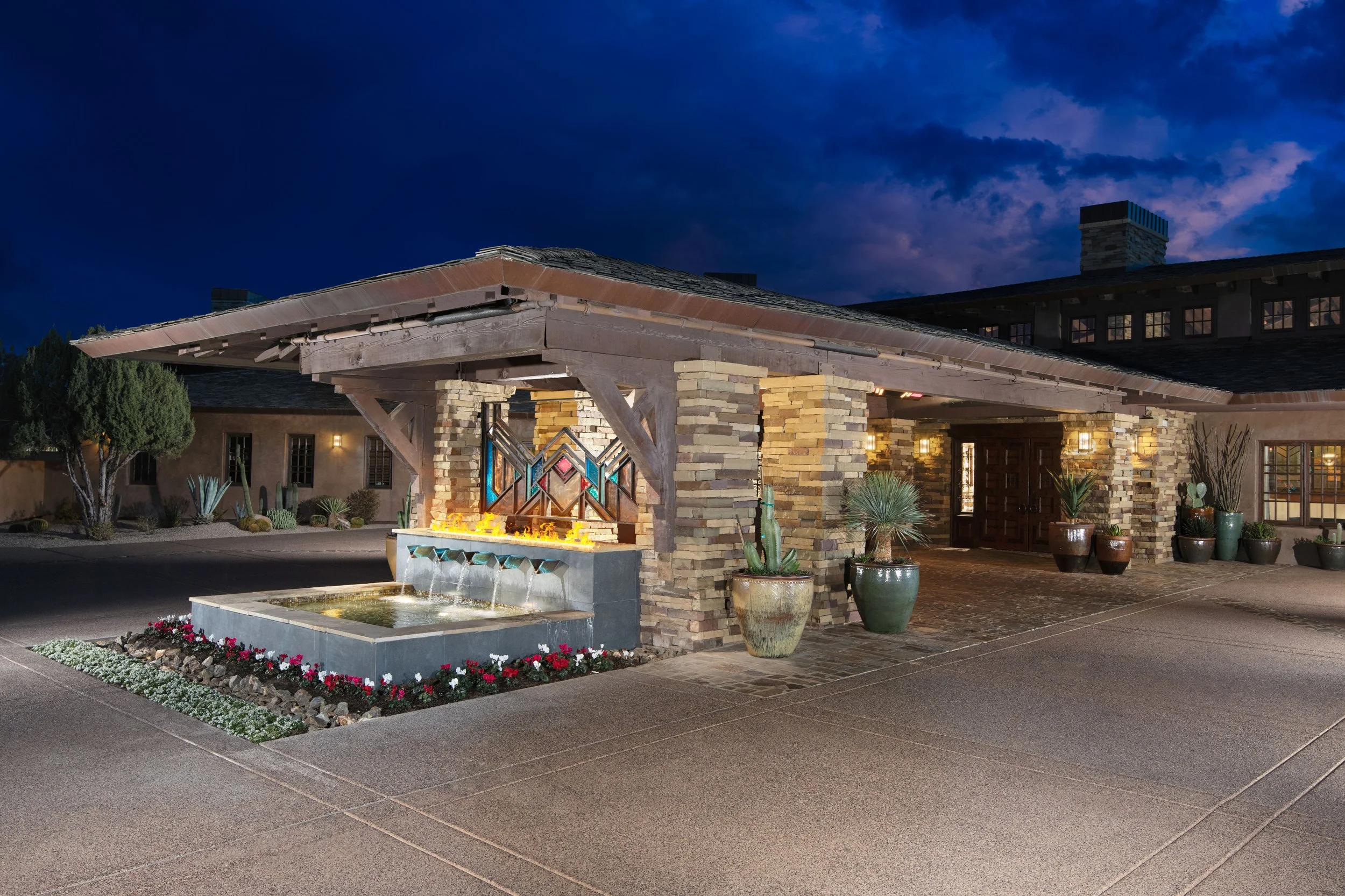 Night view of a building entrance with a porch, stone pillars, decorative glass window, potted plants, and a water fountain with small flowers in front.