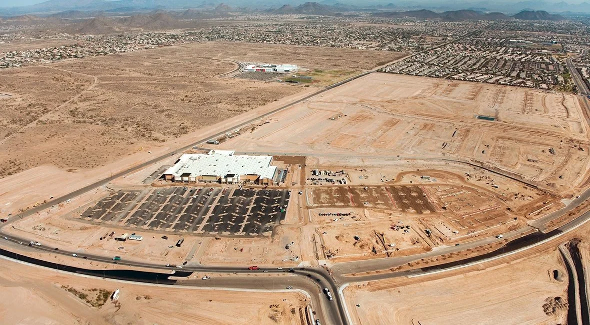 An aerial view of a large construction site in a desert area with an adjacent parking lot and a warehouse building, with a cityscape and mountains in the background.