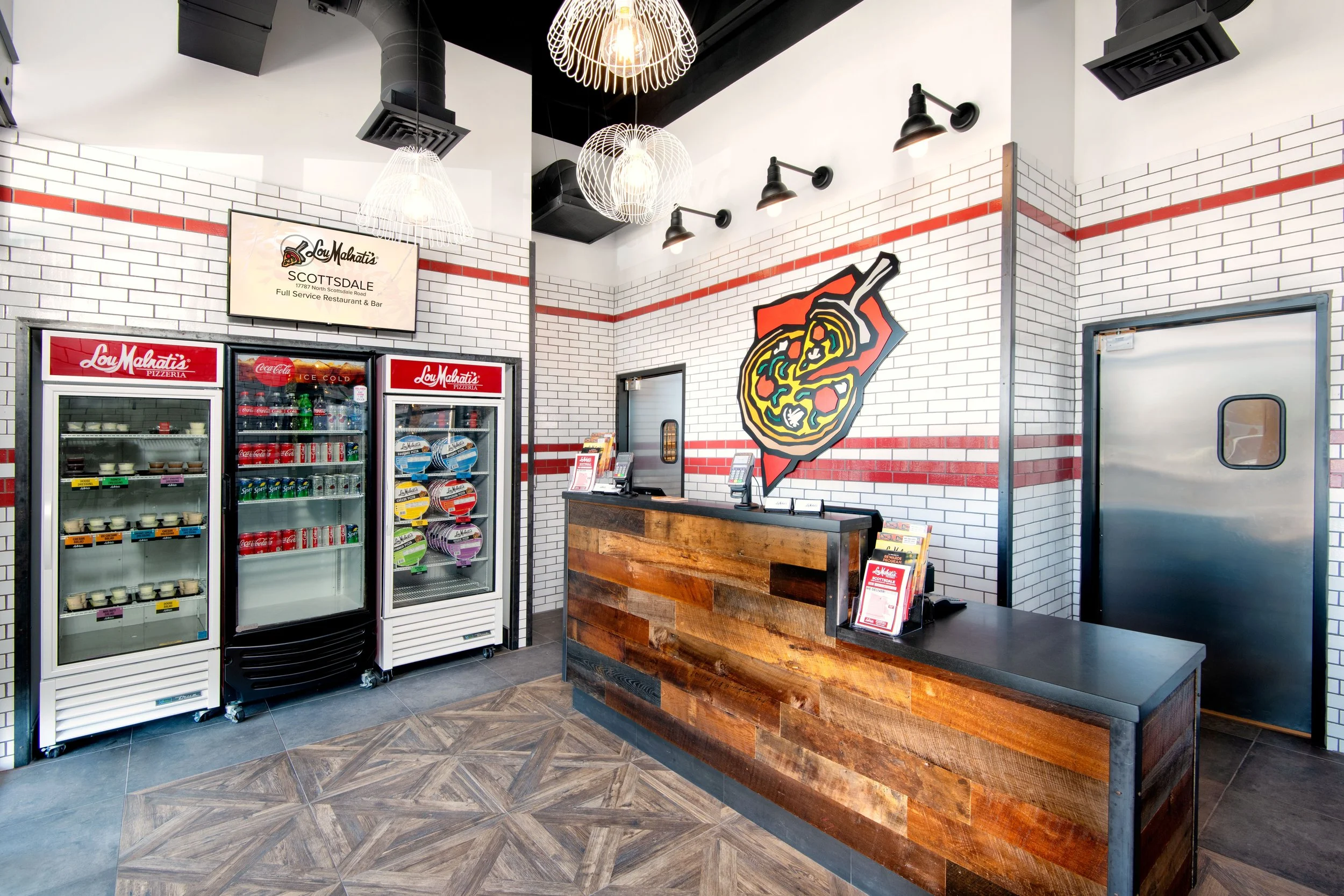 Interior of a pizzeria with a wooden counter, a seating area, and wall decor including a large pizza mural. Refrigerators with frozen foods and drinks are on the left. The walls are white with red brick accents, and the ceiling has black ducts and mo
