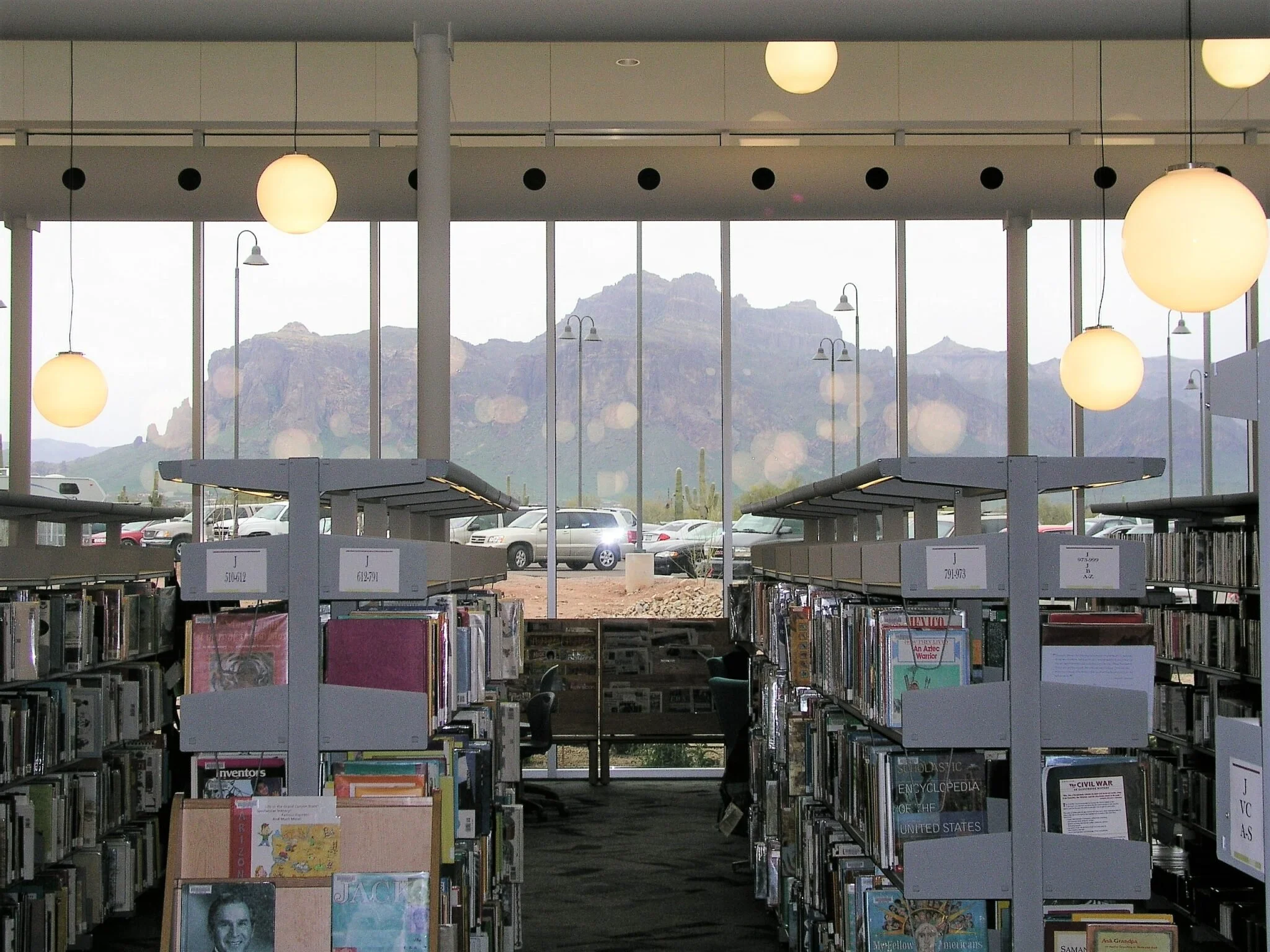 Interior of a library with bookshelves, large windows showing mountain scenery, and hanging globe lights.