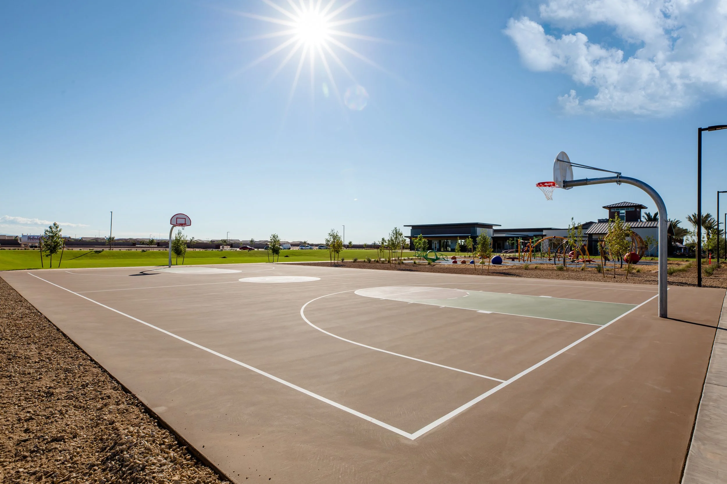 Empty outdoor basketball court with two hoops and a playground in the background under a bright sunny sky.