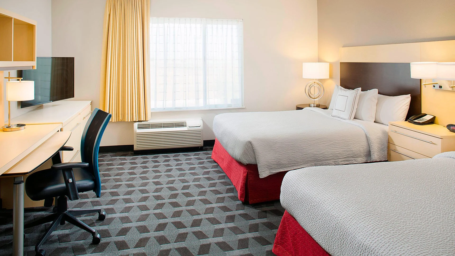 A hotel room with two beds, a desk with a chair, a mounted flat-screen TV, a window with curtains, and a nightstand with a telephone.
