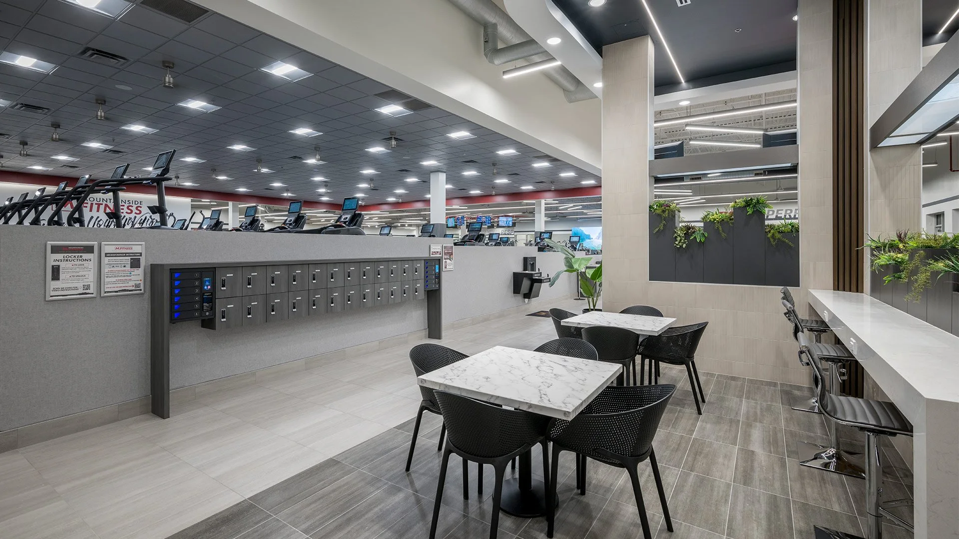A gym lobby with lockers, a seating area with marble tables and black chairs, and a counter with bar stools. The gym area has treadmills and cardio equipment, with bright ceiling lights and modern decor.