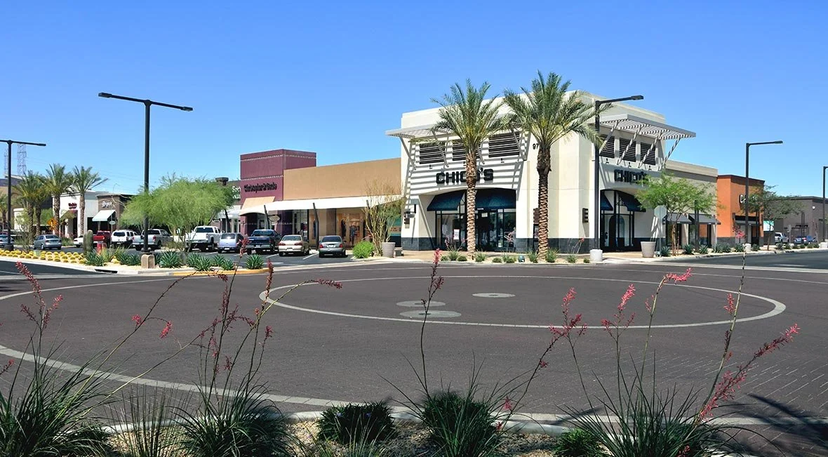 Outdoor scene of a shopping center with stores, parking lot, palm trees, and clear blue sky.