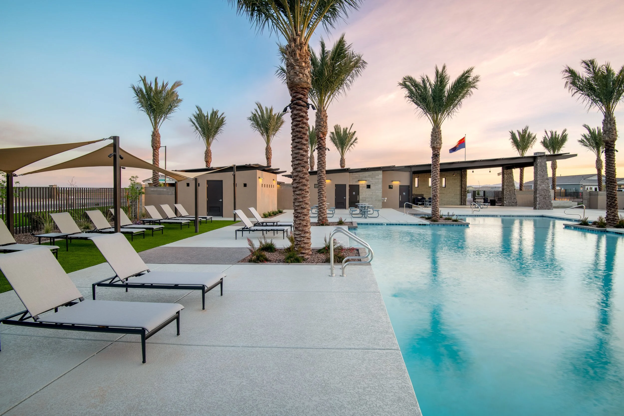 An outdoor swimming pool area with lounge chairs, palm trees, and a clubhouse at sunset.