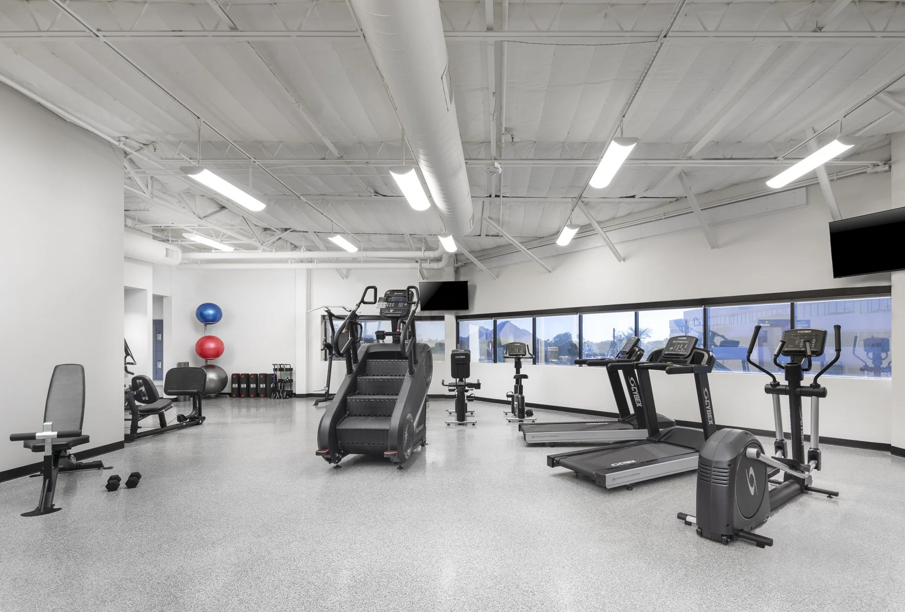 Empty gym with exercise machines, treadmill, stationary bike, elliptical, and fitness balls, in a bright space with windows and ceiling lights.