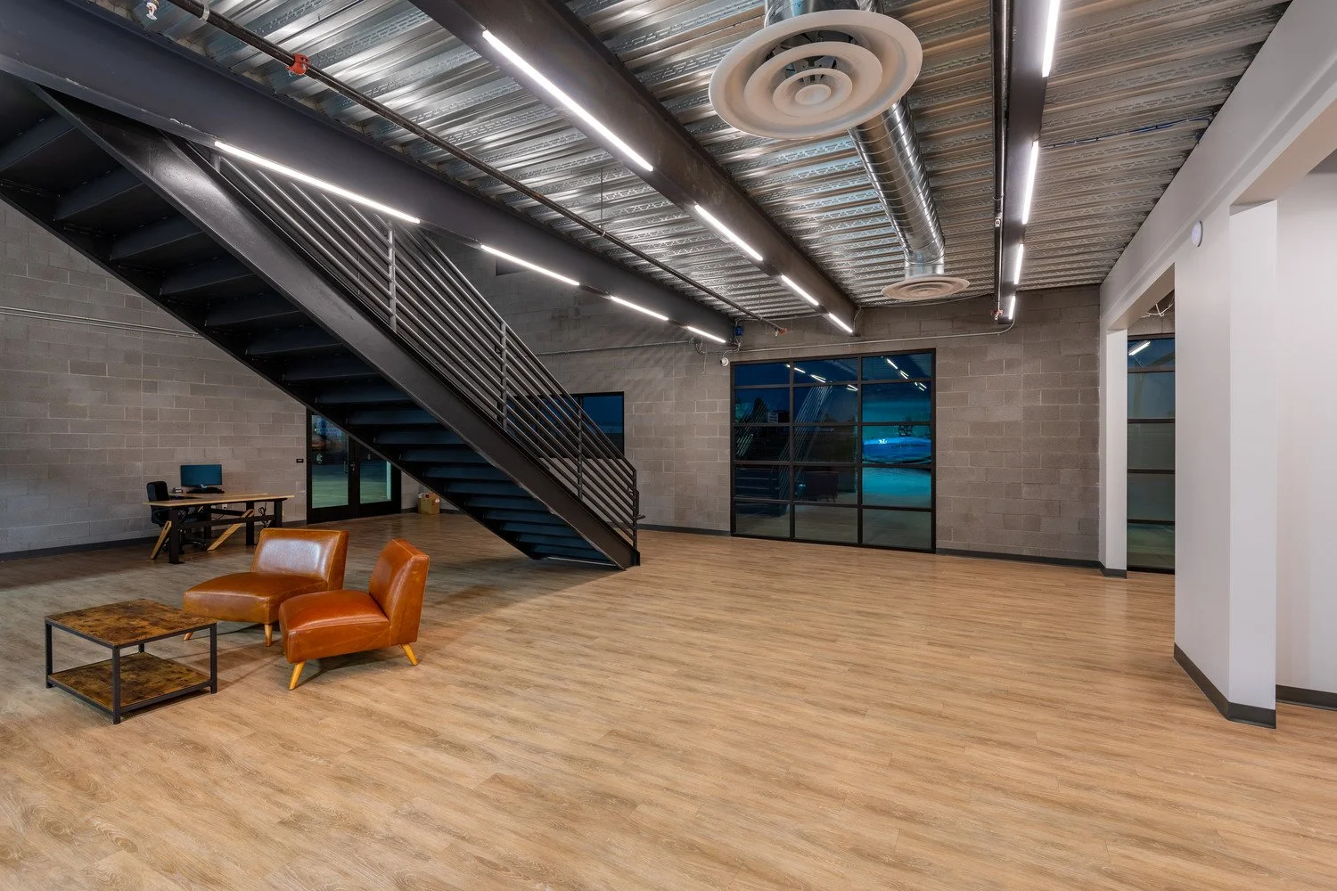 Empty industrial-style office space with wooden flooring, a staircase with black railings, large windows, and a seating area with two orange chairs and a small wooden coffee table.