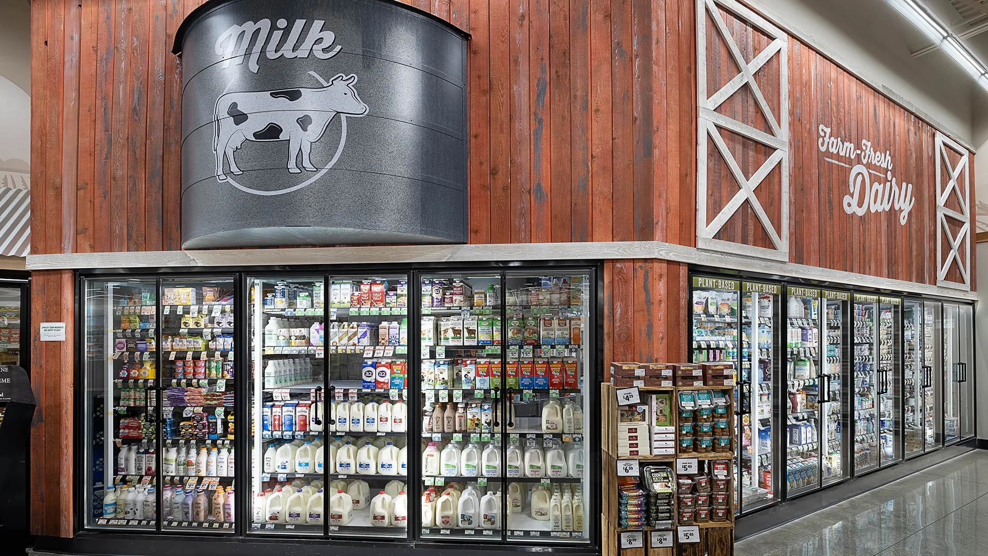 All-natural dairy products in refrigerated display case at a farm-themed grocery store section with a barn-themed sign reading 'Farm-Fresh Dairy' and a cow logo.