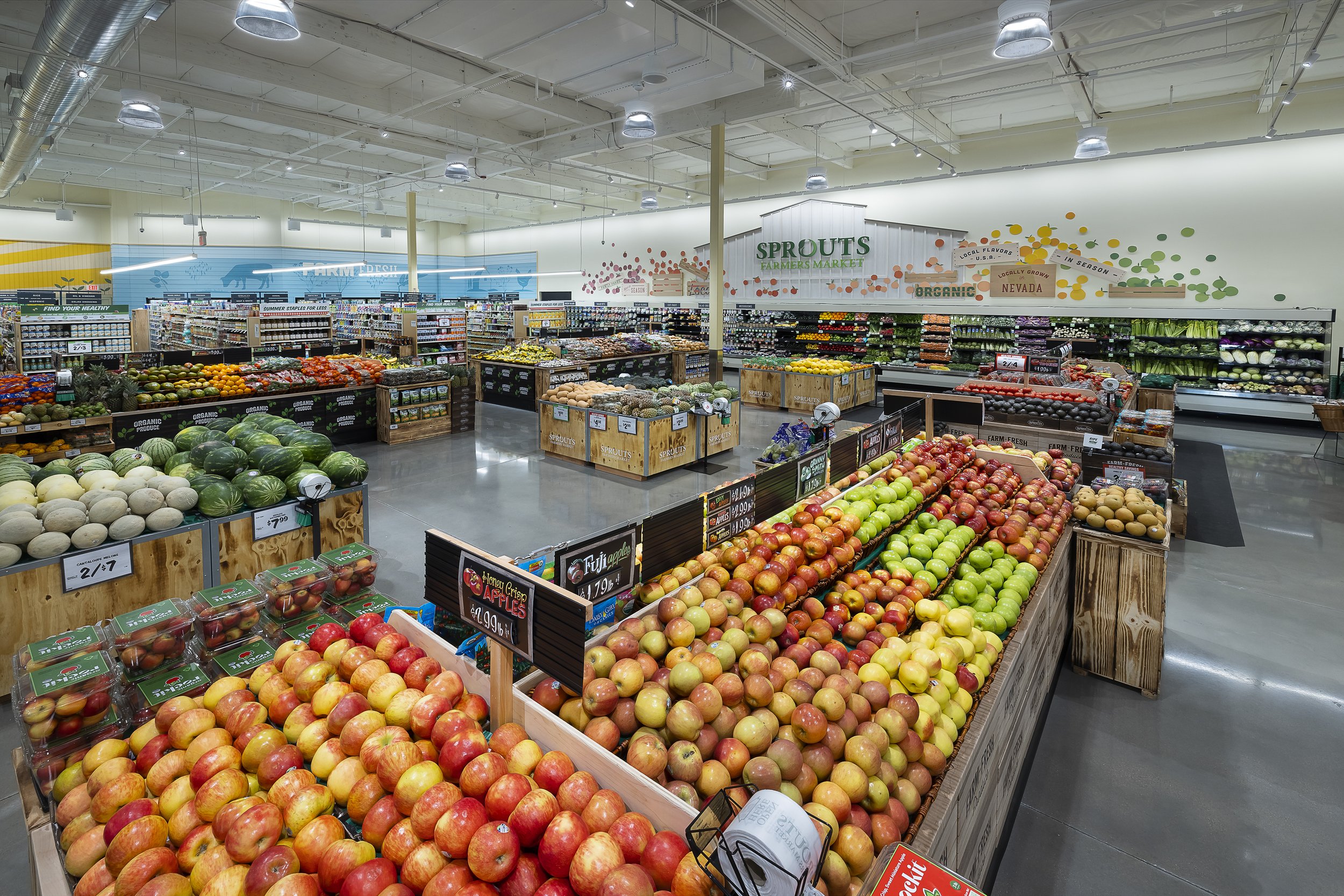 Interior of a grocery store with display bins of apples, watermelons, melons, and vegetables, with signage for organic produce and a sign reading 'Sprouts Farmers Market' in the background.