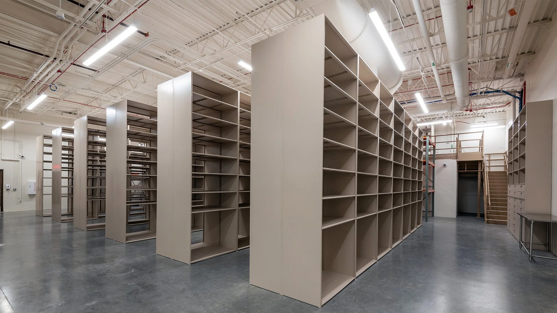 Empty industrial shelving units in large, well-lit warehouse space with concrete floor and exposed ceiling pipes.