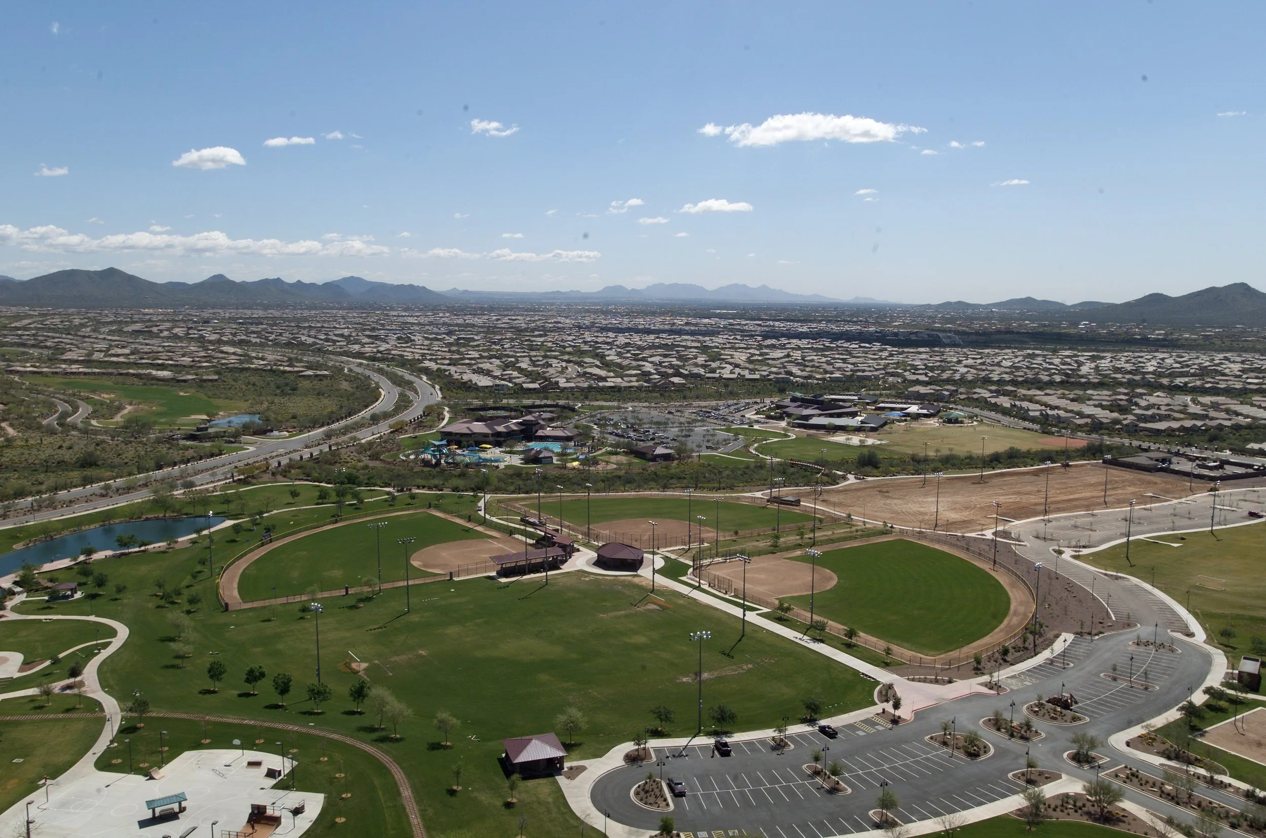 An aerial view of a large park with baseball fields, parking lots, and surrounding residential area under a clear blue sky.