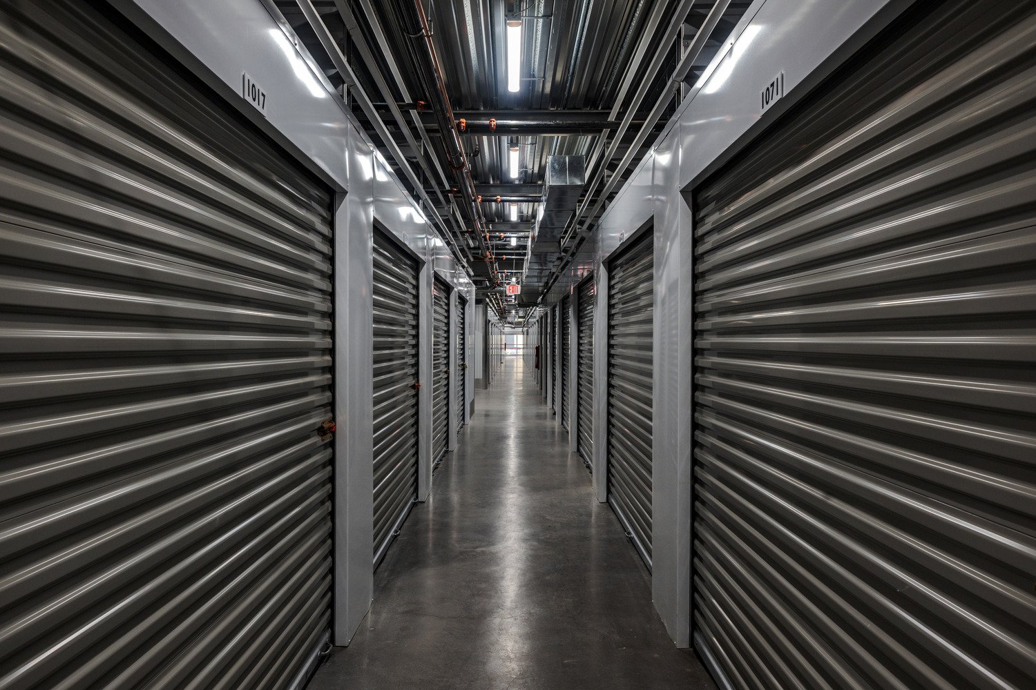 Long hallway in a storage facility with metal roll-up storage unit doors on both sides, number labels on doors, industrial lighting, and visible piping and ductwork on the ceiling.