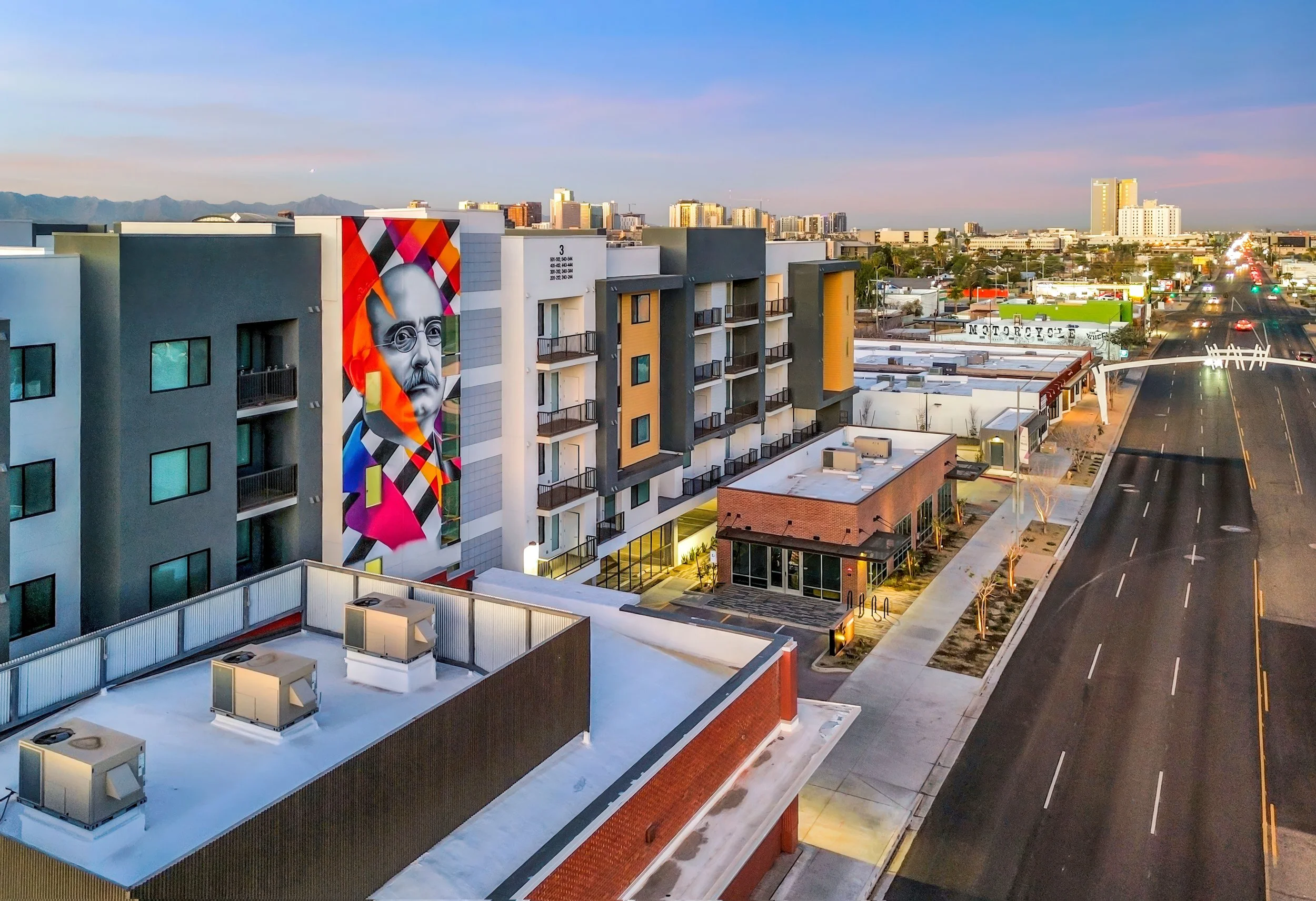 An aerial view of a city street with apartment buildings, a colorful mural of a man's face on a building, and distant high-rise buildings under a clear sky at sunset.