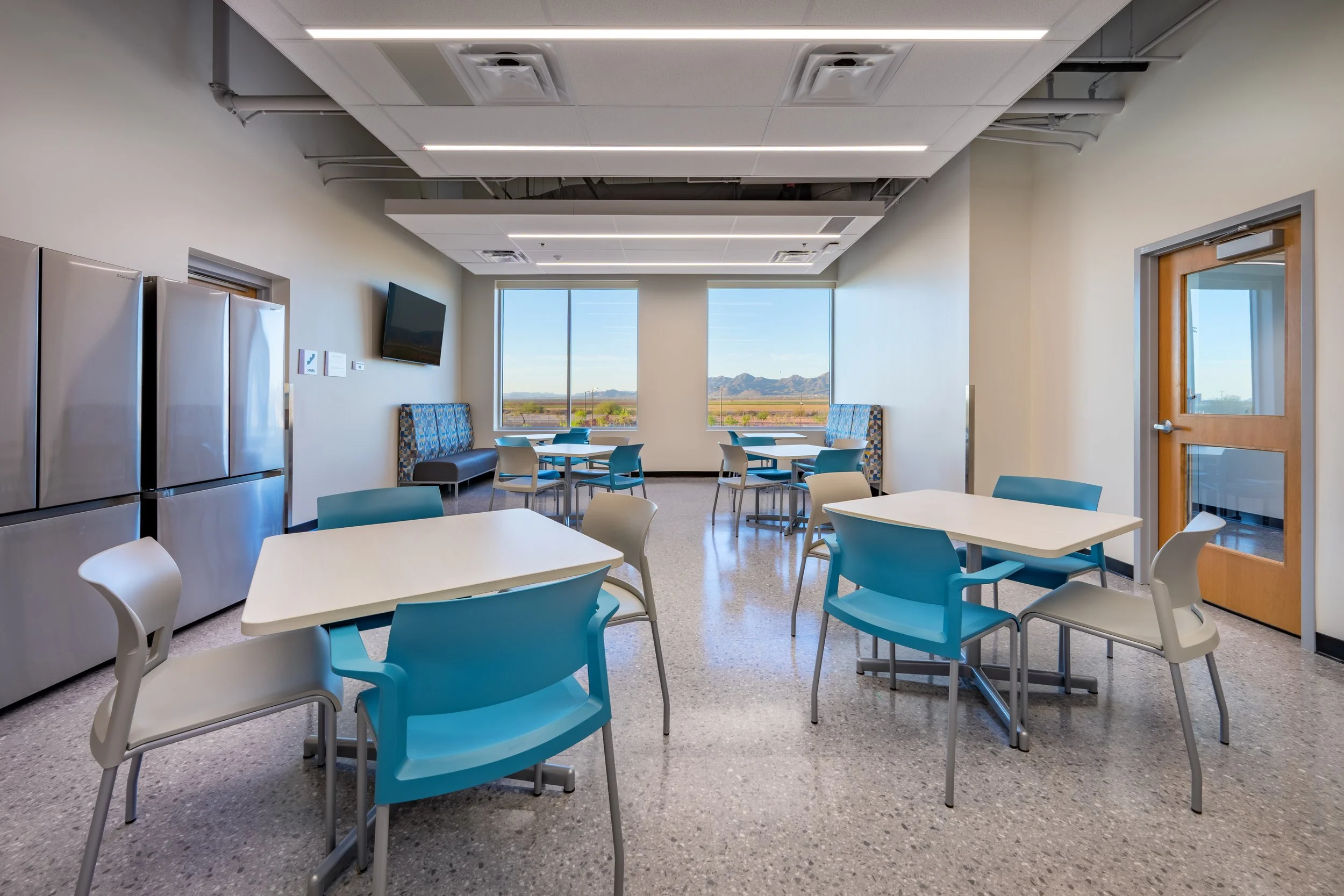 A bright, modern break room with several tables and chairs, large windows showing a mountain landscape, a wall-mounted TV, and stainless steel refrigerators.