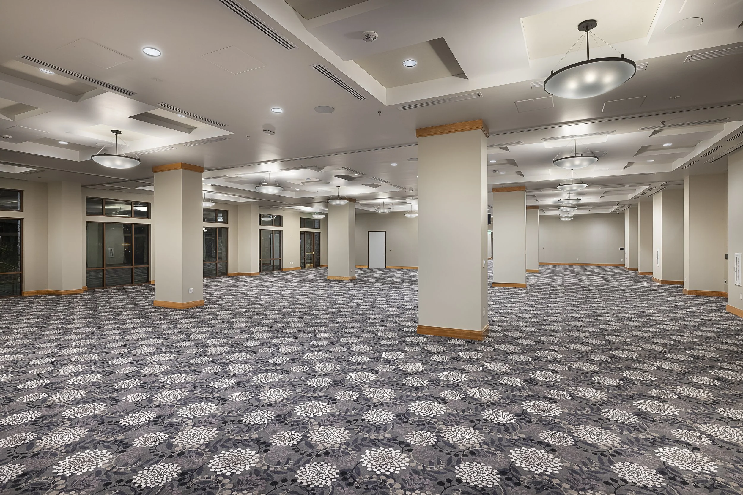 Empty indoor event space with patterned carpet, white walls, multiple ceiling lights, and large windows and doors along the left side.