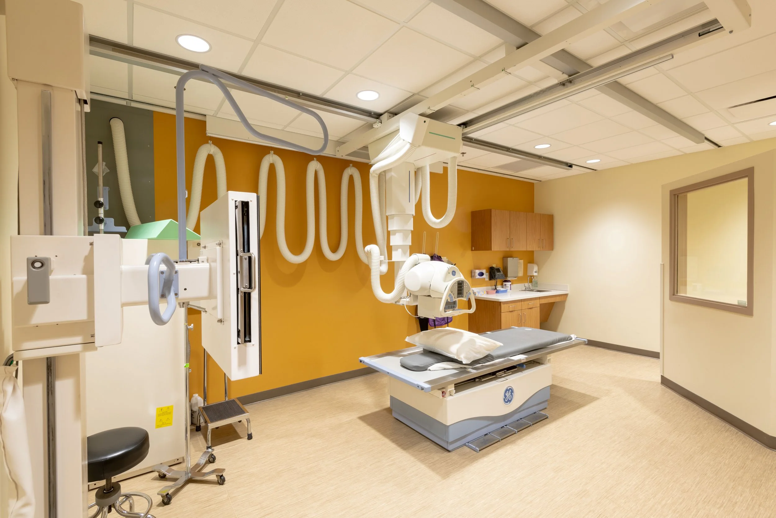 Hospital room with X-ray machine and bed, beige walls, brown accent wall, medical cabinets, and equipment.