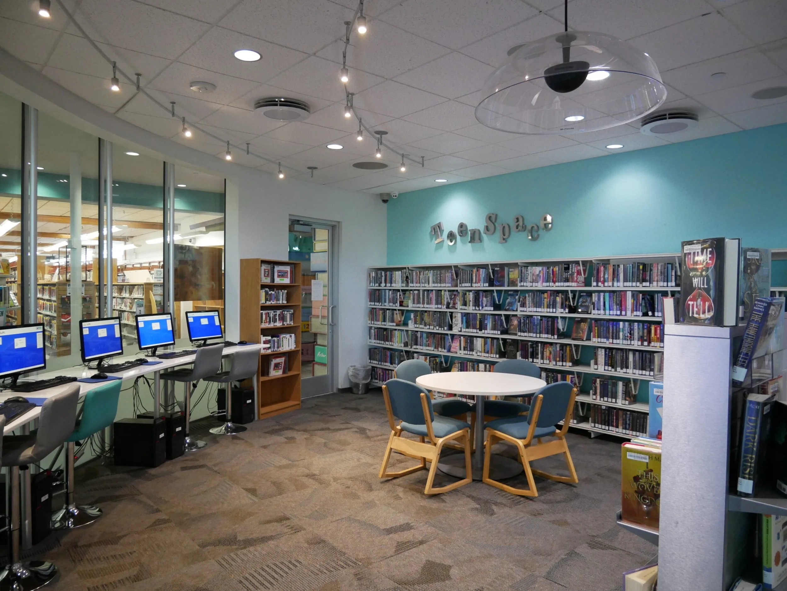 A library room with four desktop computers on a counter near the window, a round table surrounded by six blue-cushioned wooden chairs, a bookshelf with books, a blue wall with the words 'Teen Space' in decorative letters, and ceiling lights.