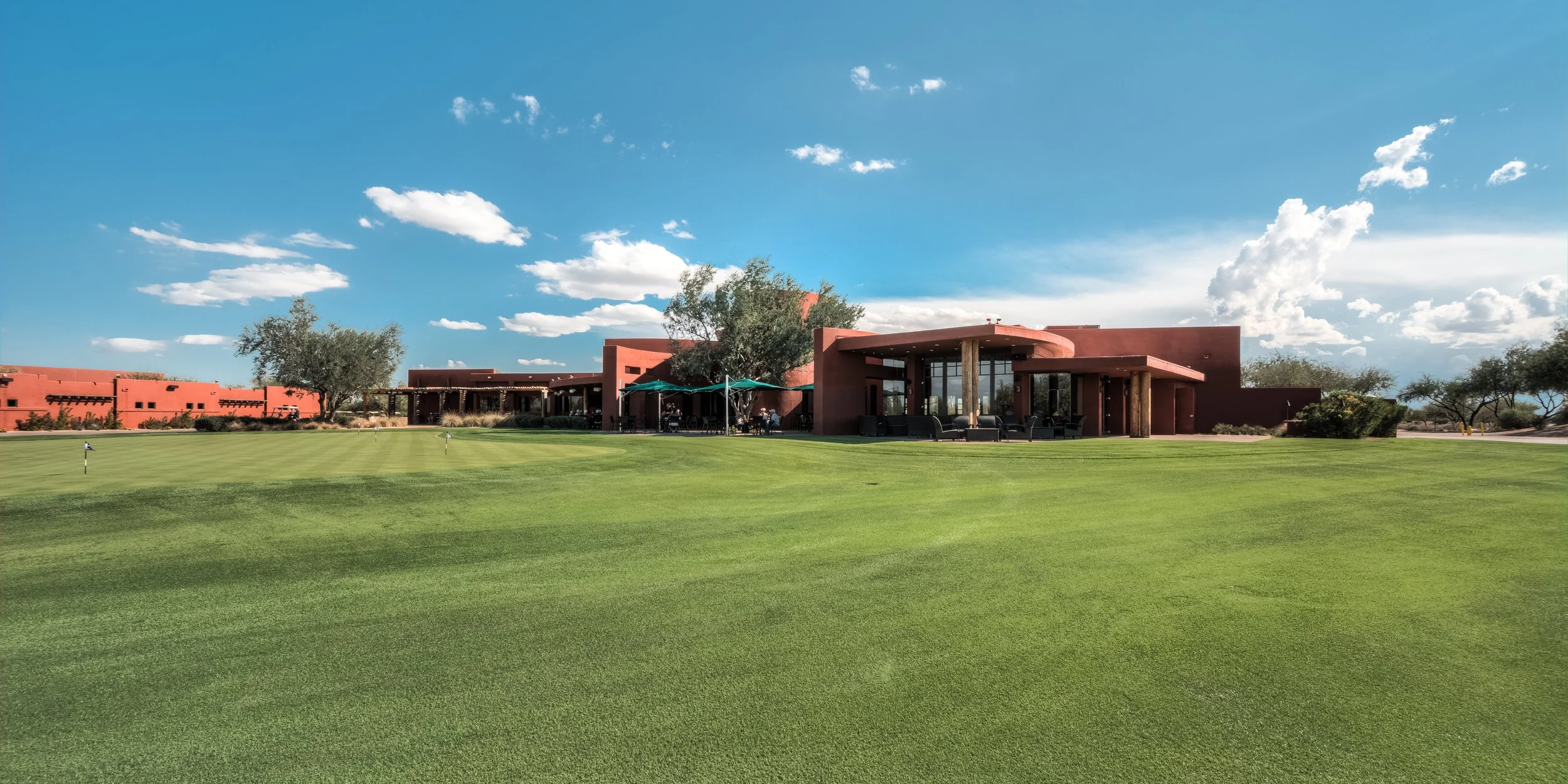 A modern building with a reddish desert-like exterior, surrounded by a well-maintained green golf course with small flags, under a partly cloudy blue sky with trees and outdoor seating areas.