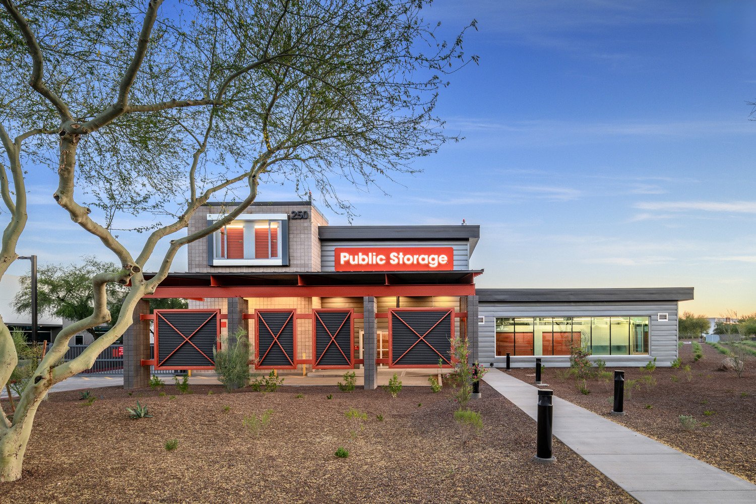 A modern public storage facility with a sign that reads 'Public Storage' in bright red letters. The building features a gray exterior with black and red accents, surrounded by a sidewalk and landscaped yard with small bushes and a large tree. The sky