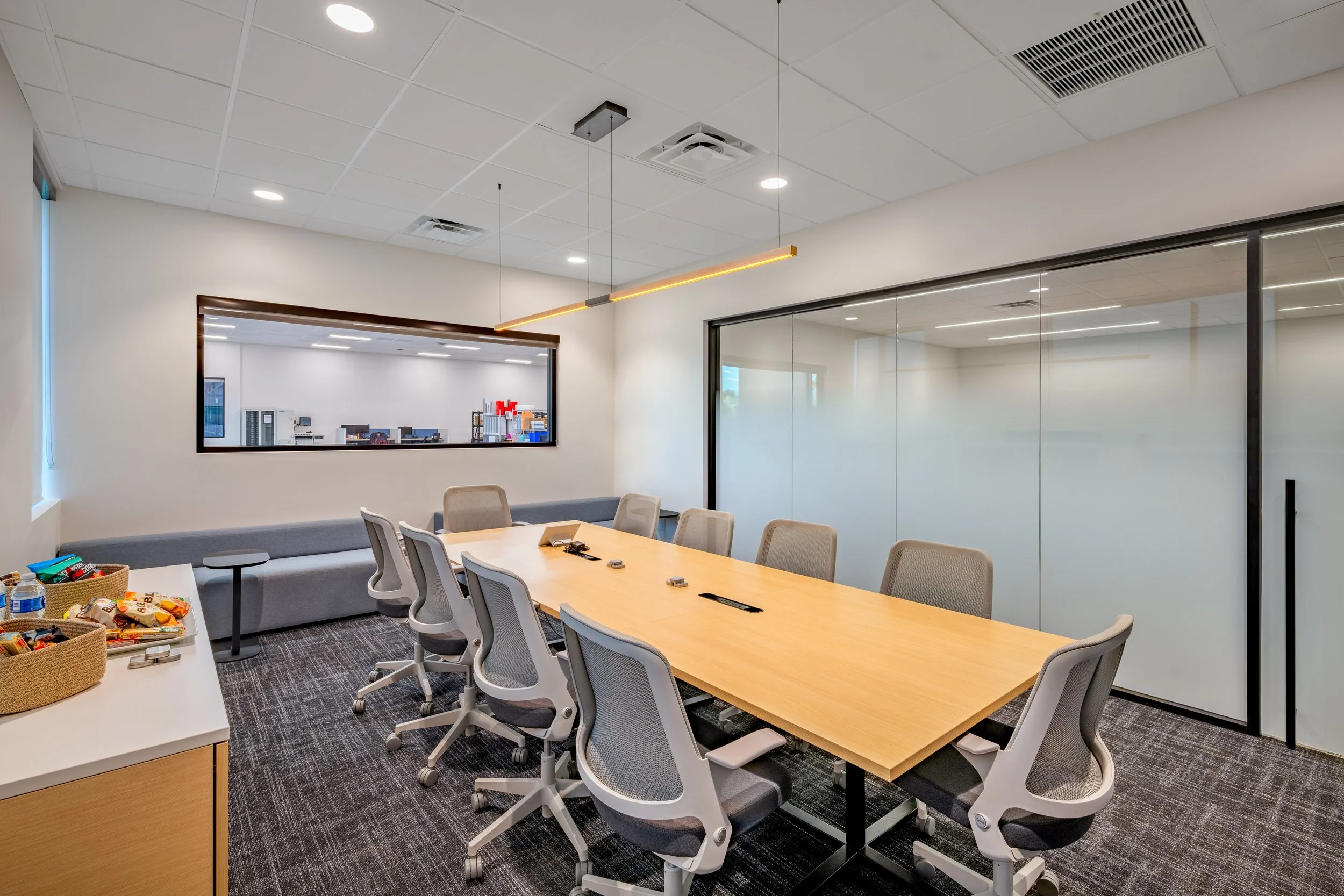 Empty modern conference room with a long wooden table, nine gray office chairs, a side table with snacks, glass wall, and a view into another room with office supplies.