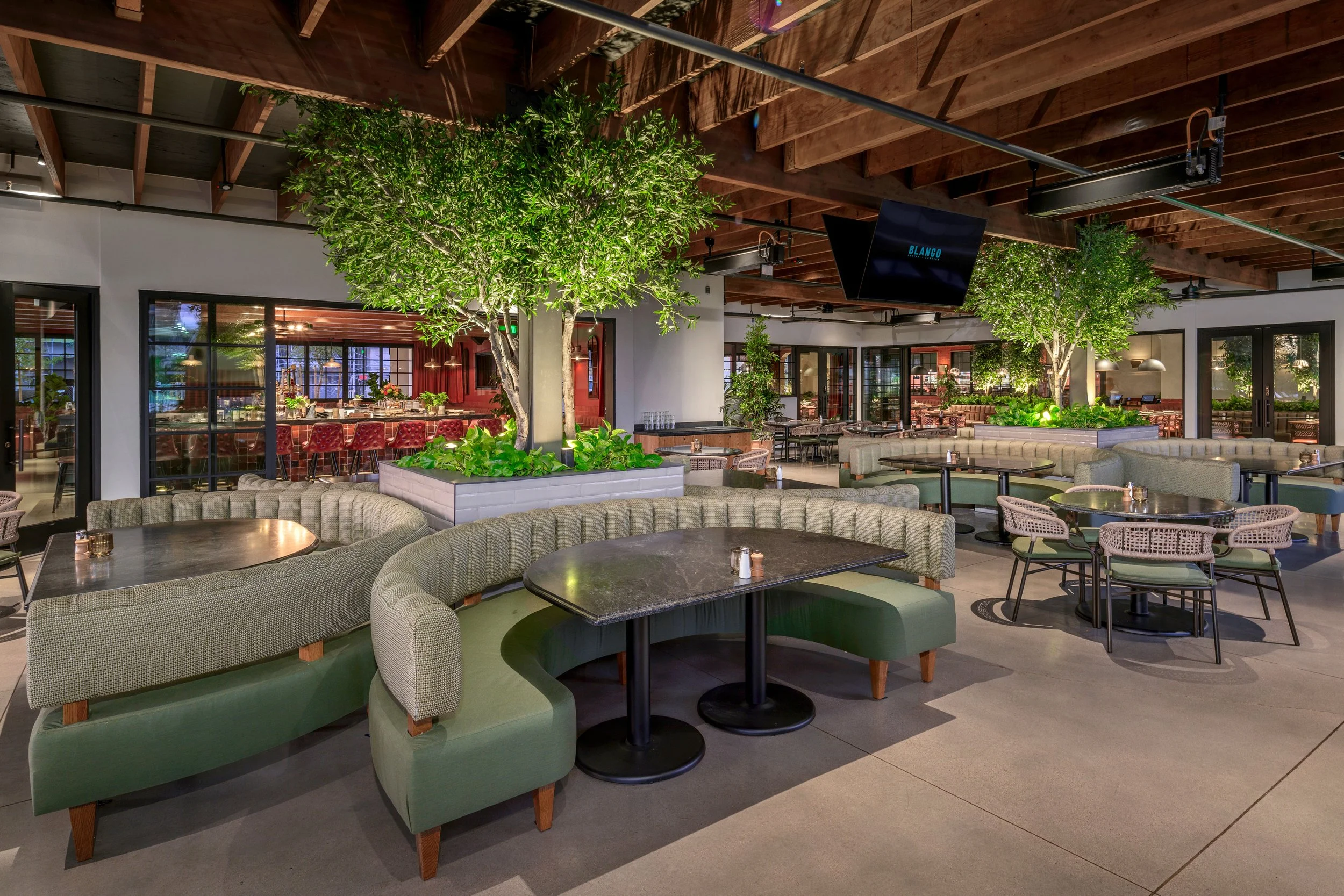 Interior of a modern restaurant or lounge with green curved booths, round and rectangular tables, indoor trees, and large glass windows.