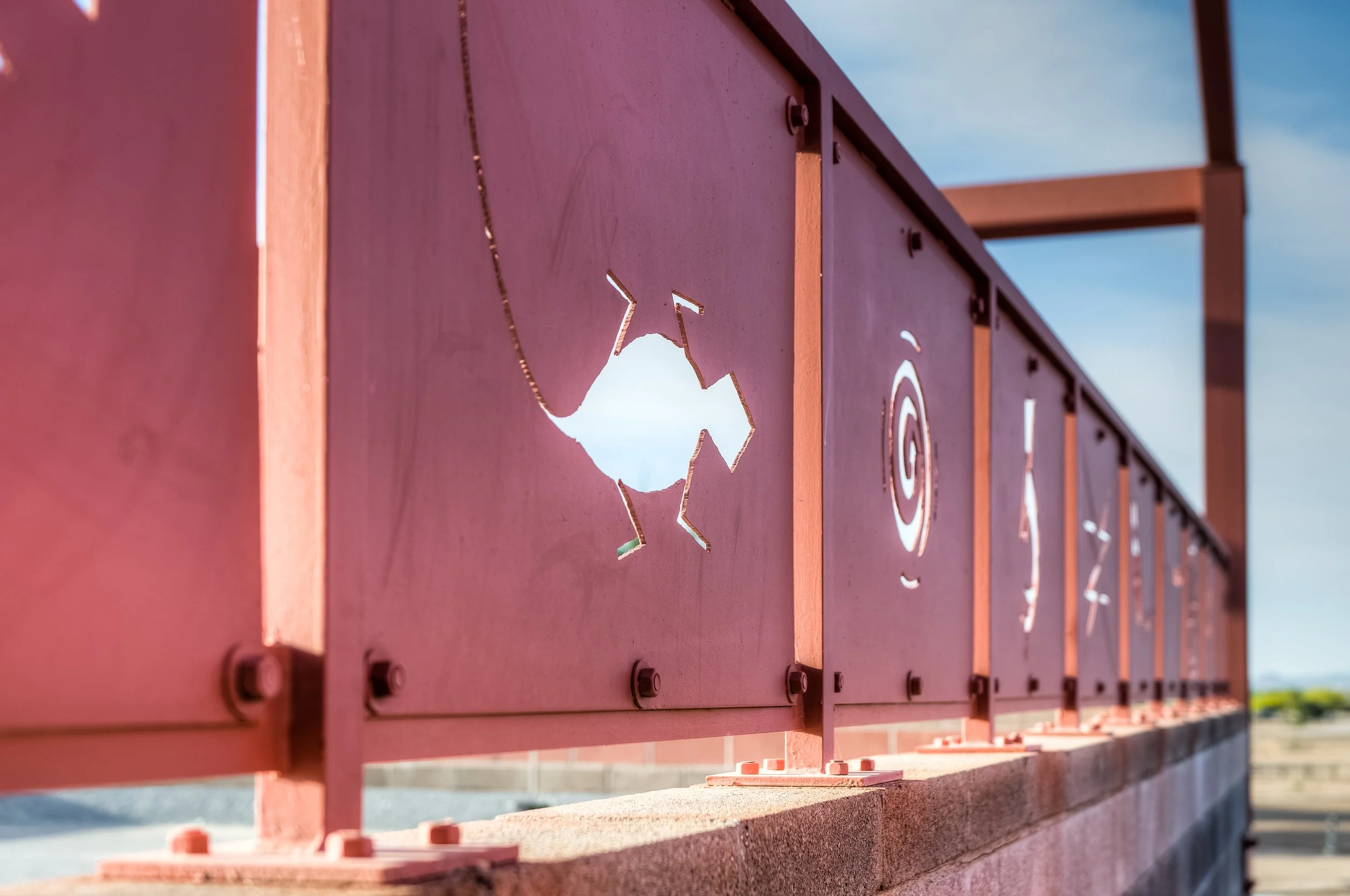 Close-up of metal panels on a structure, each panel cut with different native american symbols, and other designs, attached to a brick base under a blue sky.