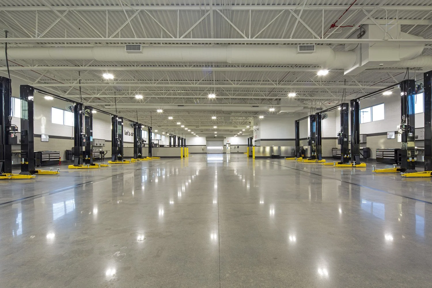 Empty vehicle service workshop with multiple lift stations lined up along the walls, bright lighting, polished concrete floors, and a corrugated metal ceiling.