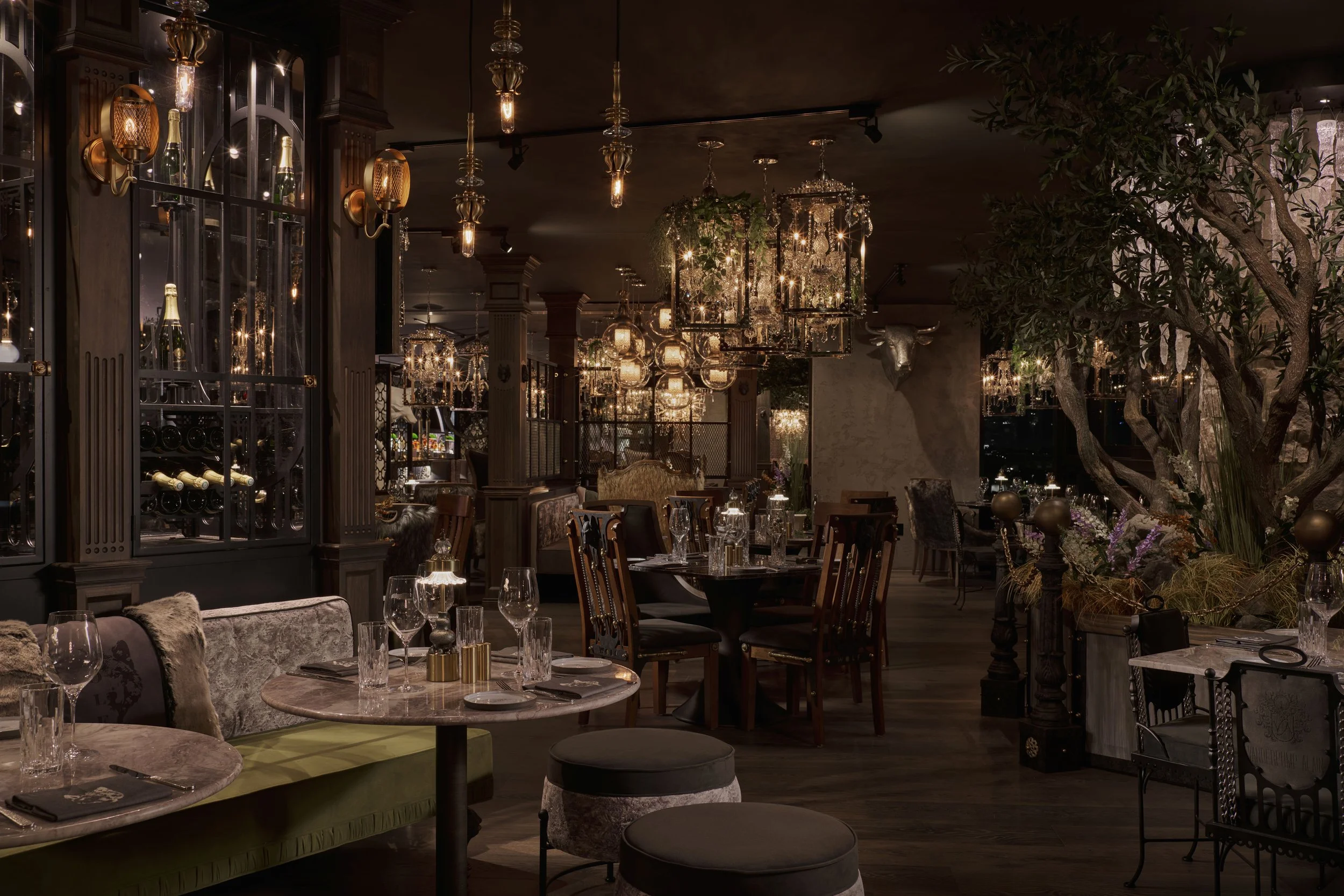 Elegant restaurant interior with dim lighting, multiple chandeliers, a large decorative tree, and well-set tables with glasses and napkins.