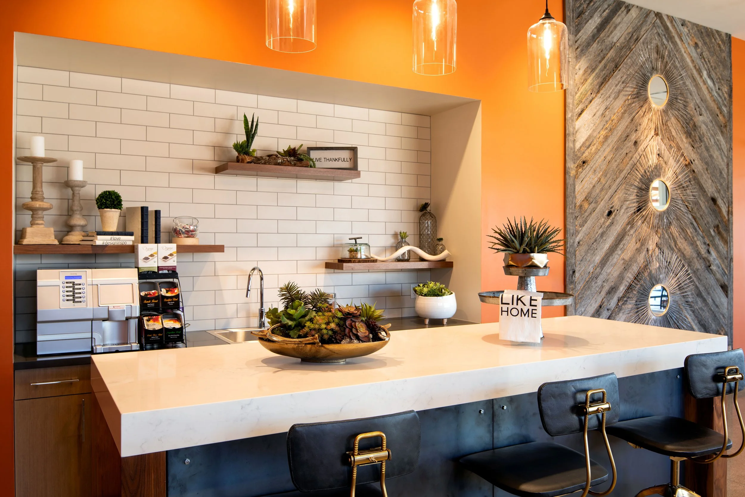 Modern kitchen with a white marble countertop, black stools, and a mix of white and wooden decor elements. Potted plants, a coffee machine, and decorative items adorn the space, with an accent wall featuring wood and mirror details.