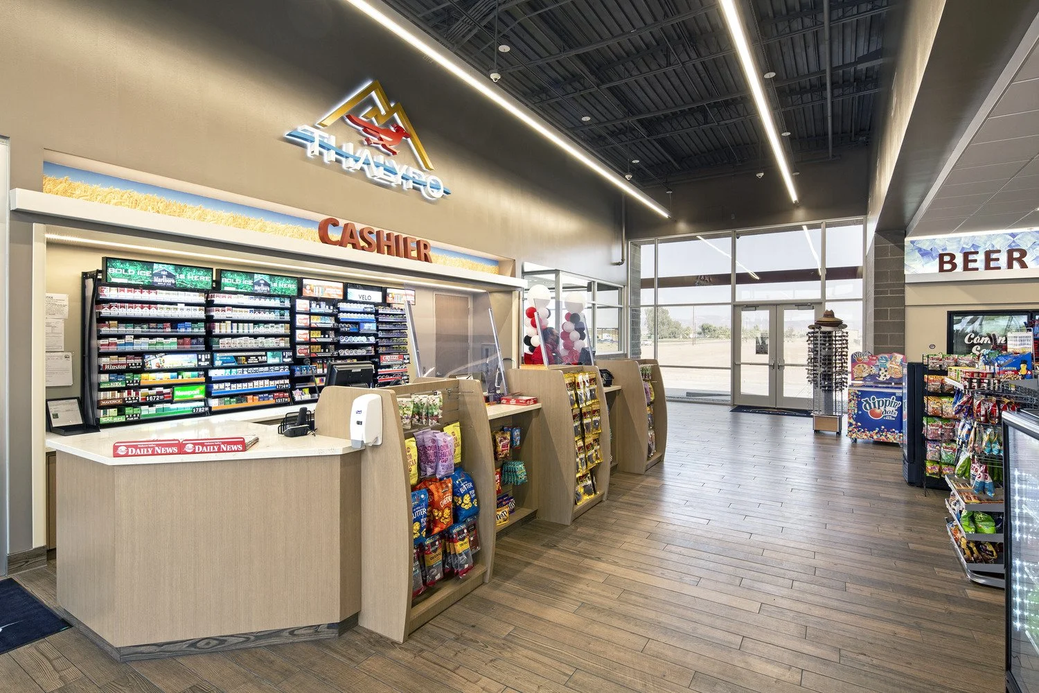 Inside a convenience store near a cashier counter with snacks, balloons, and a large glass entrance door.