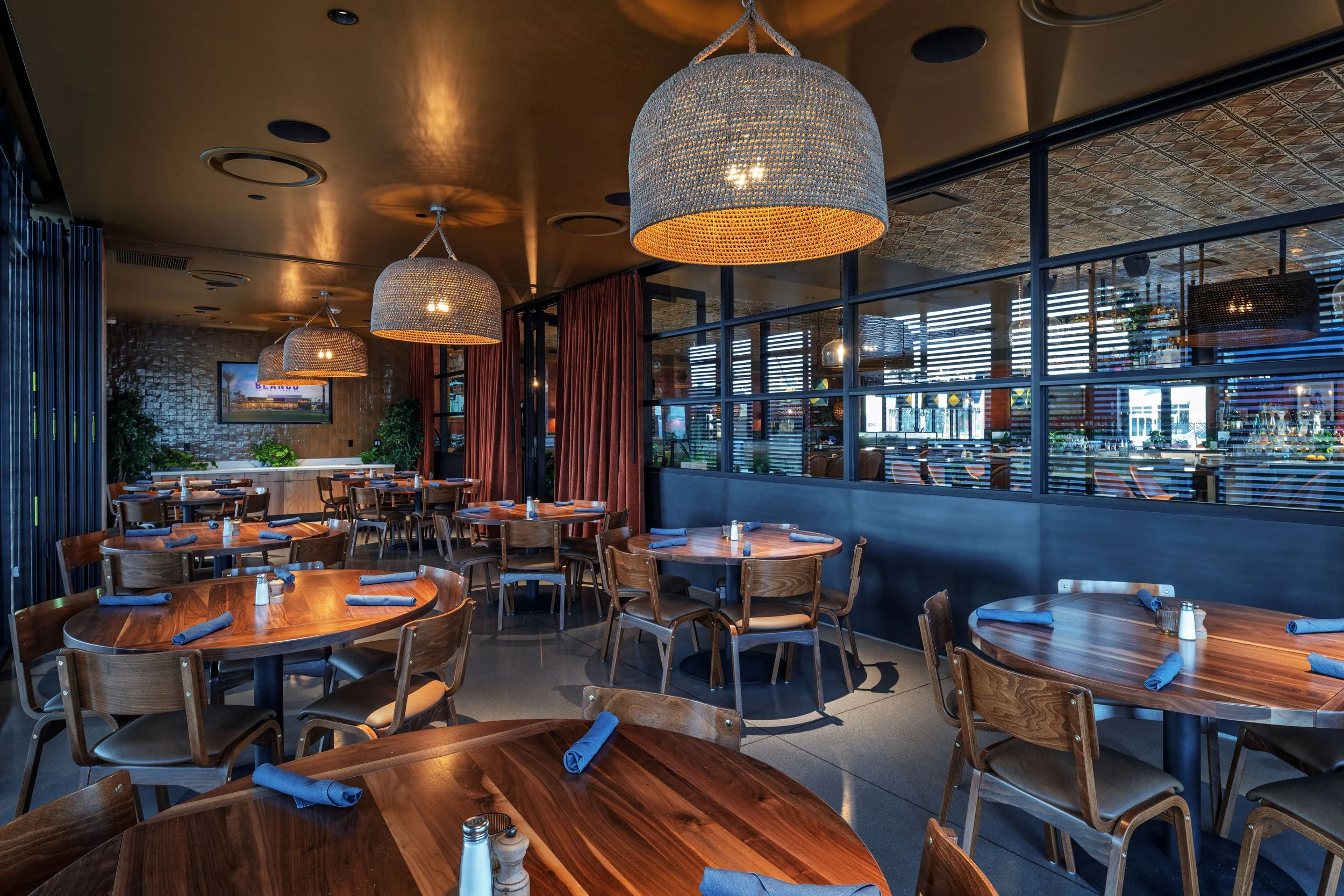 Empty restaurant with round wooden tables, wooden chairs, blue napkins, large woven hanging pendant lights, and a separated dining area with glass walls and wooden blinds.