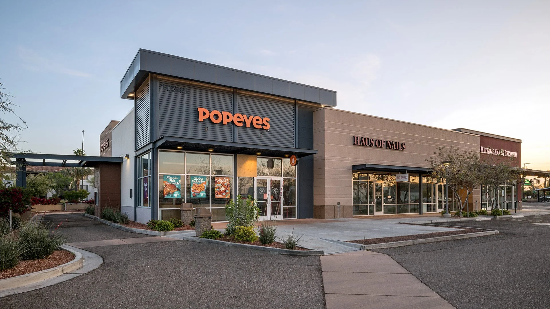 An outdoor view of a strip mall showing a Popeyes restaurant, a nail salon named Haus of Nails, and a store called Microvana & Premium, with a parking lot and landscaped areas with small shrubs and trees.