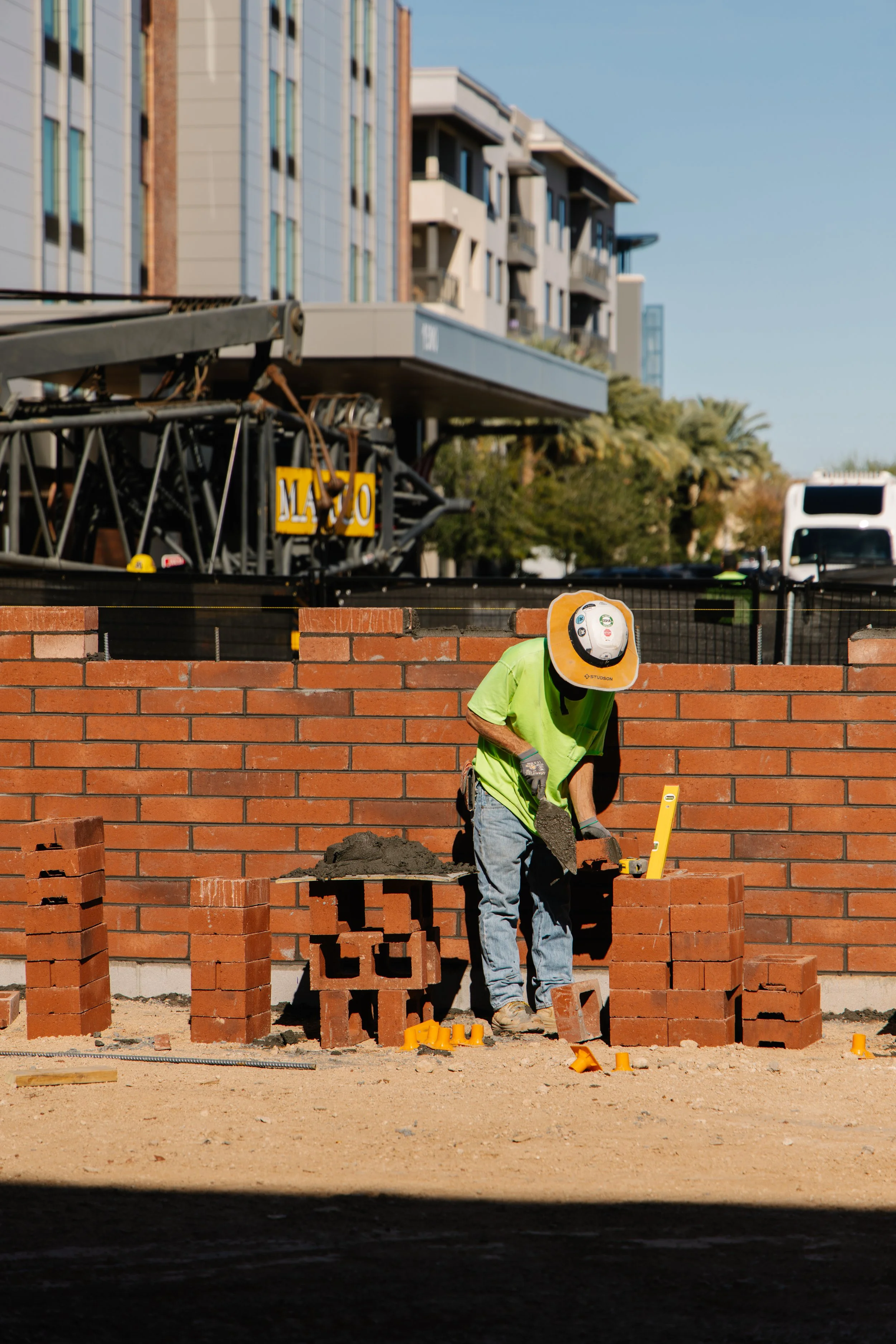 Construction worker laying bricks, wearing a green shirt and a white safety helmet, working on a brick wall building site with residential buildings in the background.