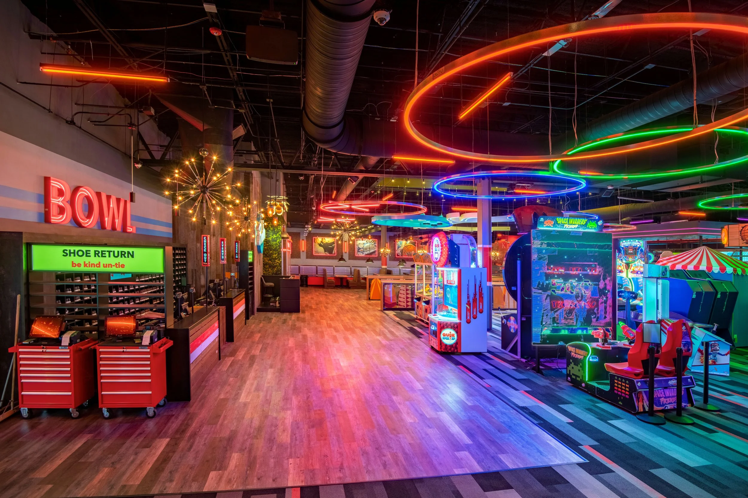 An interior arcade with neon lights, arcade machines, and a shoe return area labeled 'BOWL' and 'be kind un-tie' sign, with a colorful, vibrant atmosphere.
