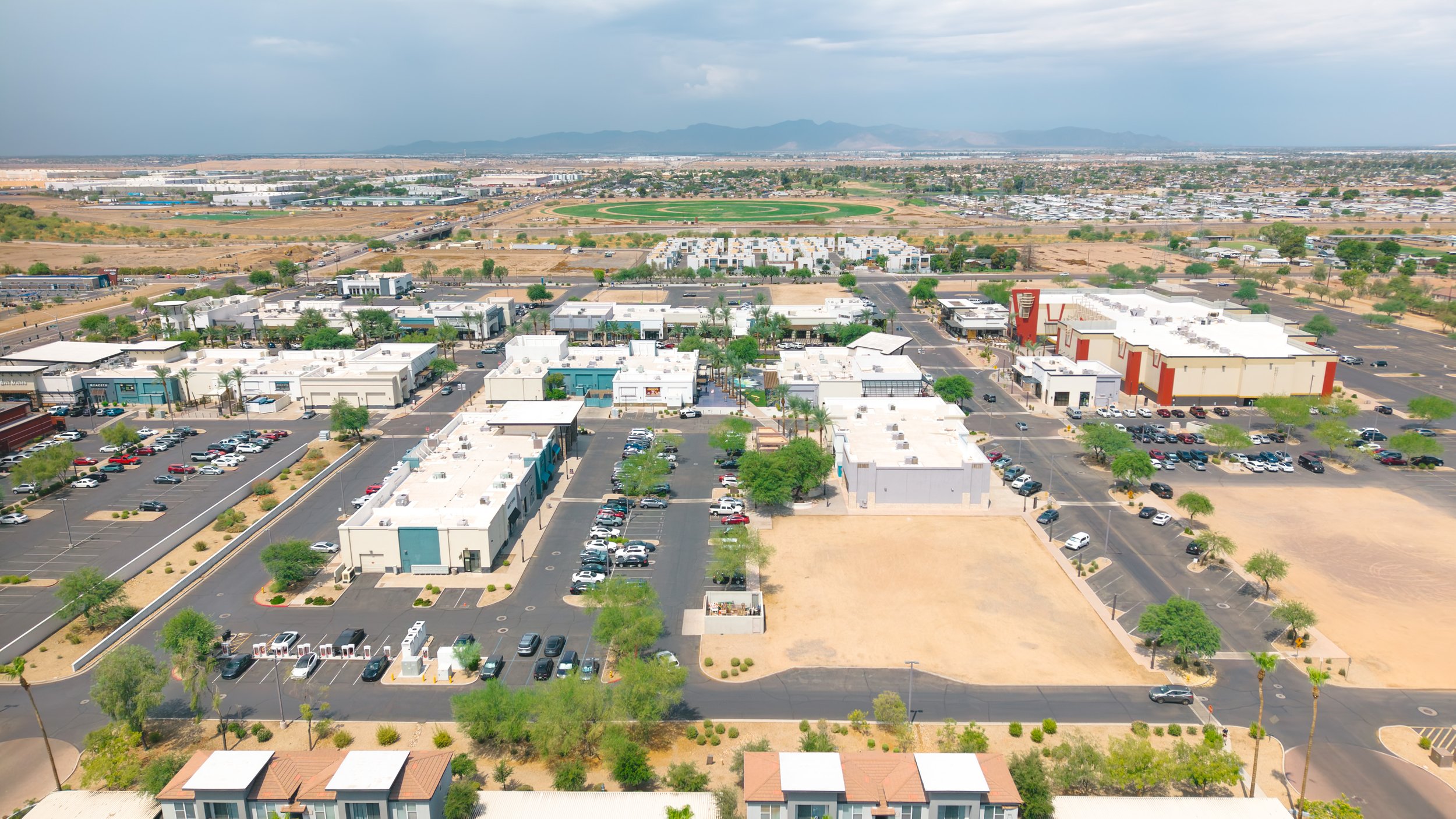 Aerial view of a commercial area with white buildings and parking lots, with a green circular park, desert landscape, and mountains in the background under a cloudy sky.