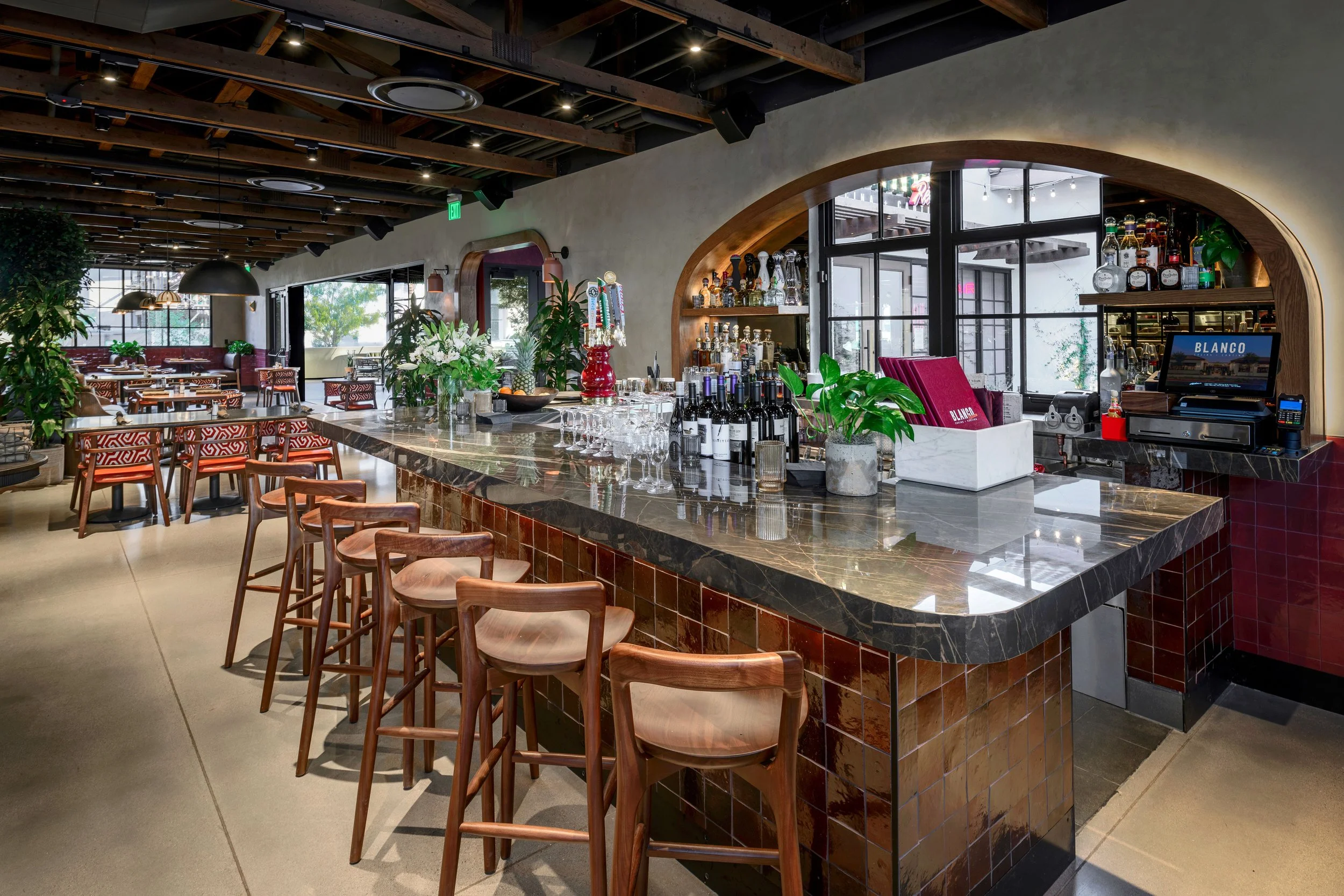 Modern restaurant bar with a marble countertop, wooden bar stools, and well-lit interior featuring plants, bottles of alcohol, and a point of sale system.