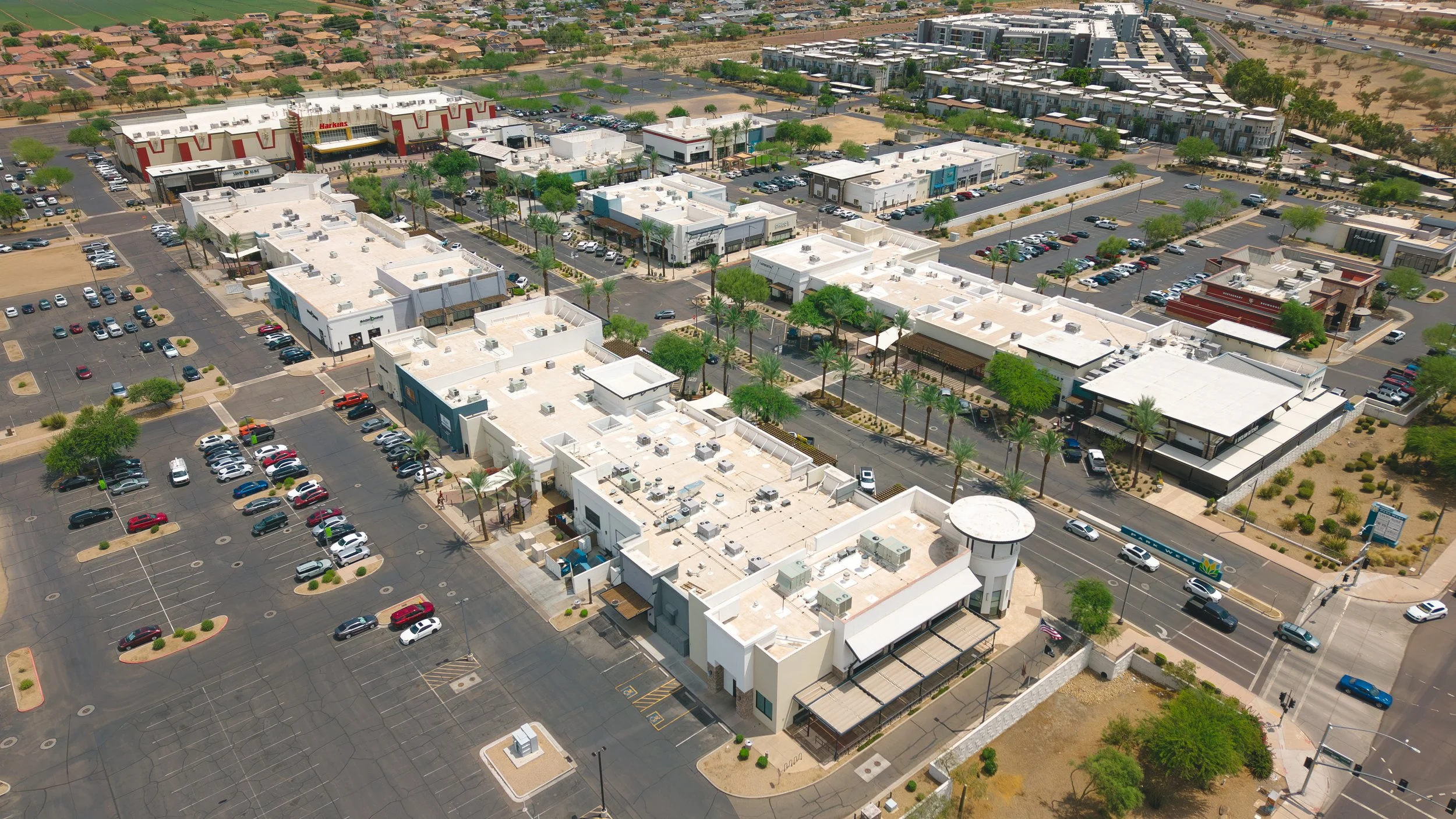 An aerial view of a shopping center with various retail stores, parking lots, and surrounding residential area with single-family homes and apartment complexes, with green trees and desert landscape.