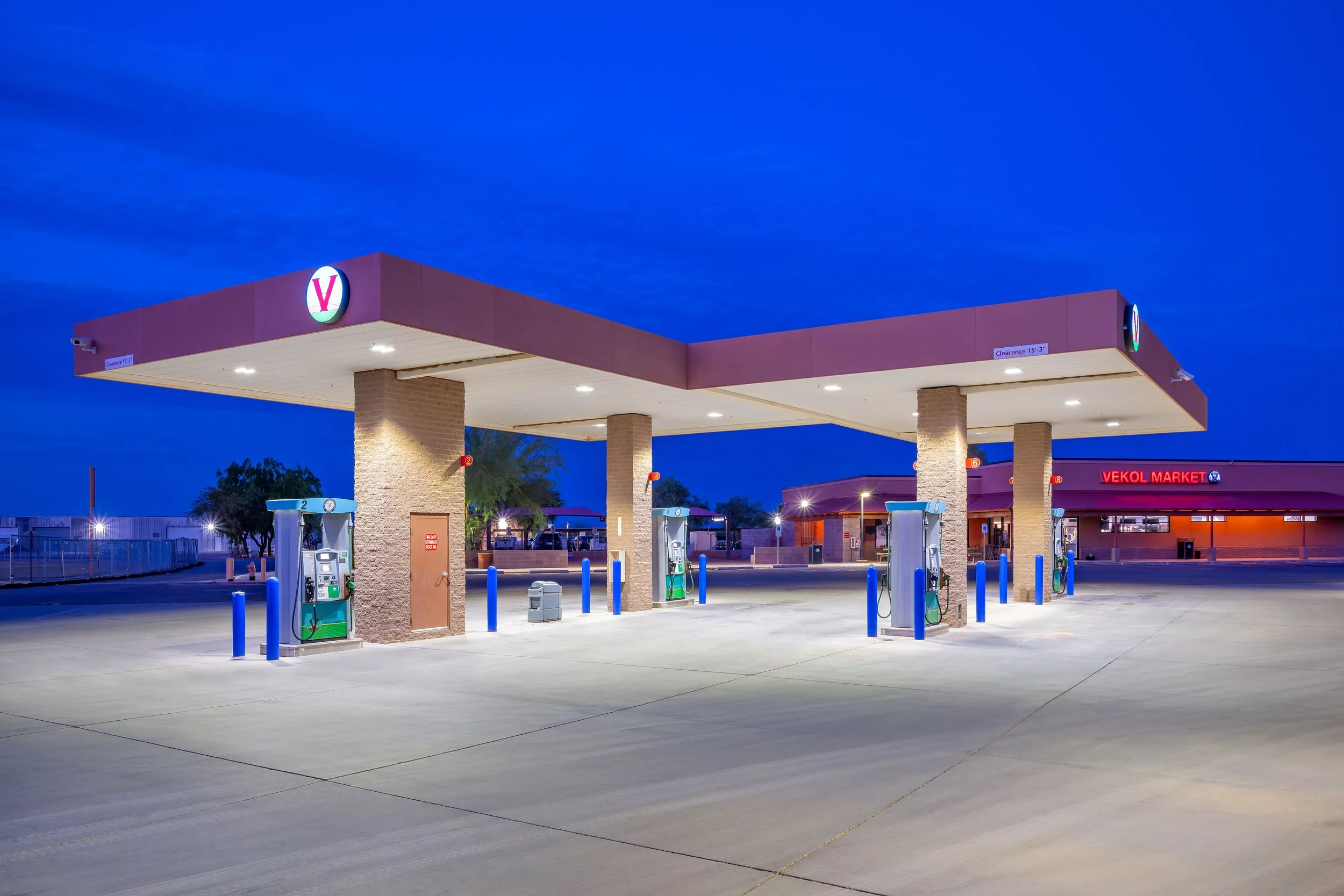Empty gas station at dusk with three fuel pumps under a canopy and a convenience store in the background.