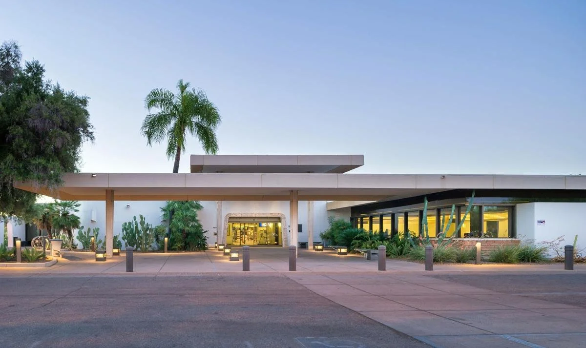 A modern building with white exterior walls, surrounded by desert plants and cactus, with a palm tree in the background. The entrance is illuminated, and the parking lot in front is bordered by short bollards.