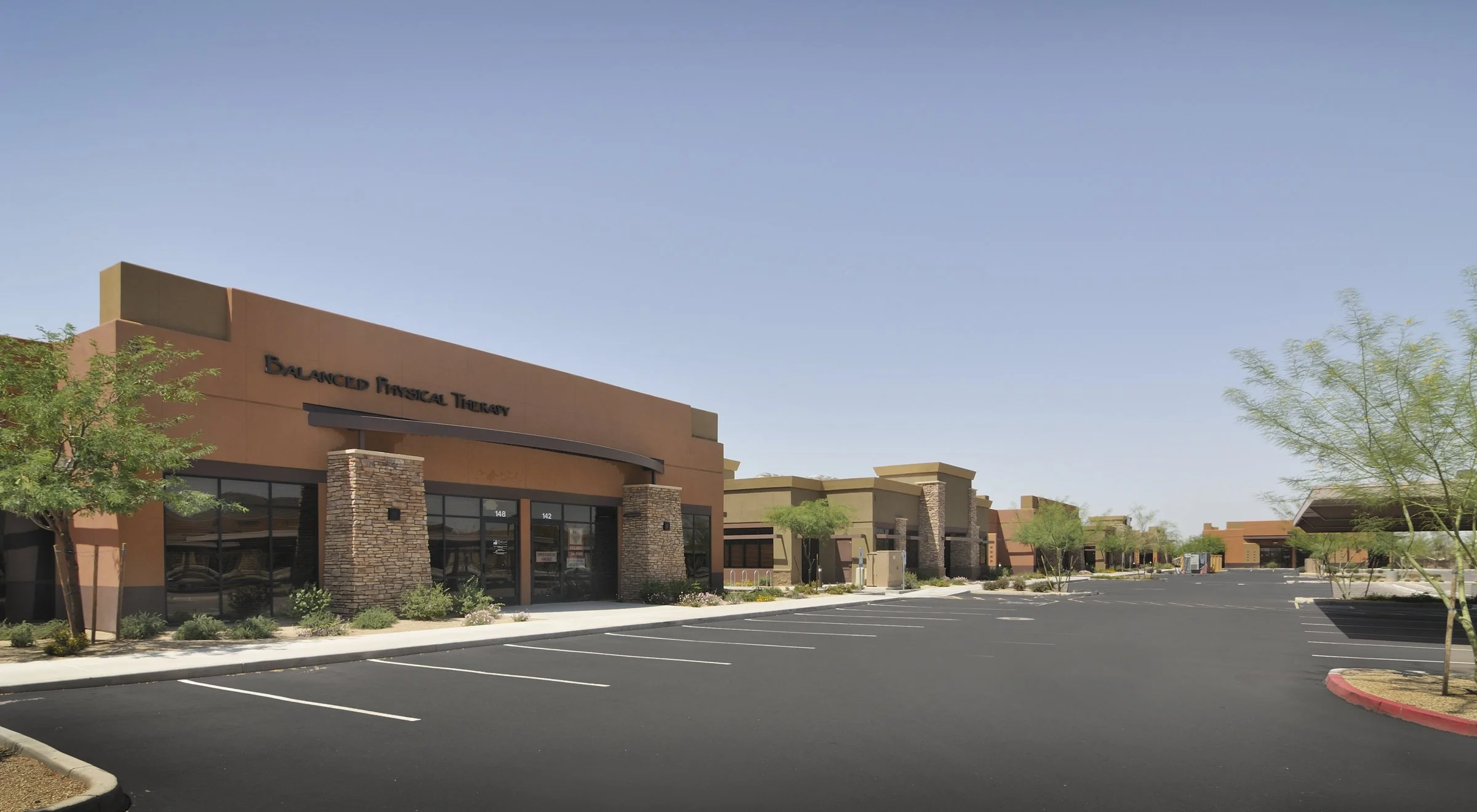 A shopping complex with a store called Balanced Physical Therapy, featuring a parking lot with marked parking spaces and a few trees, under a clear blue sky.
